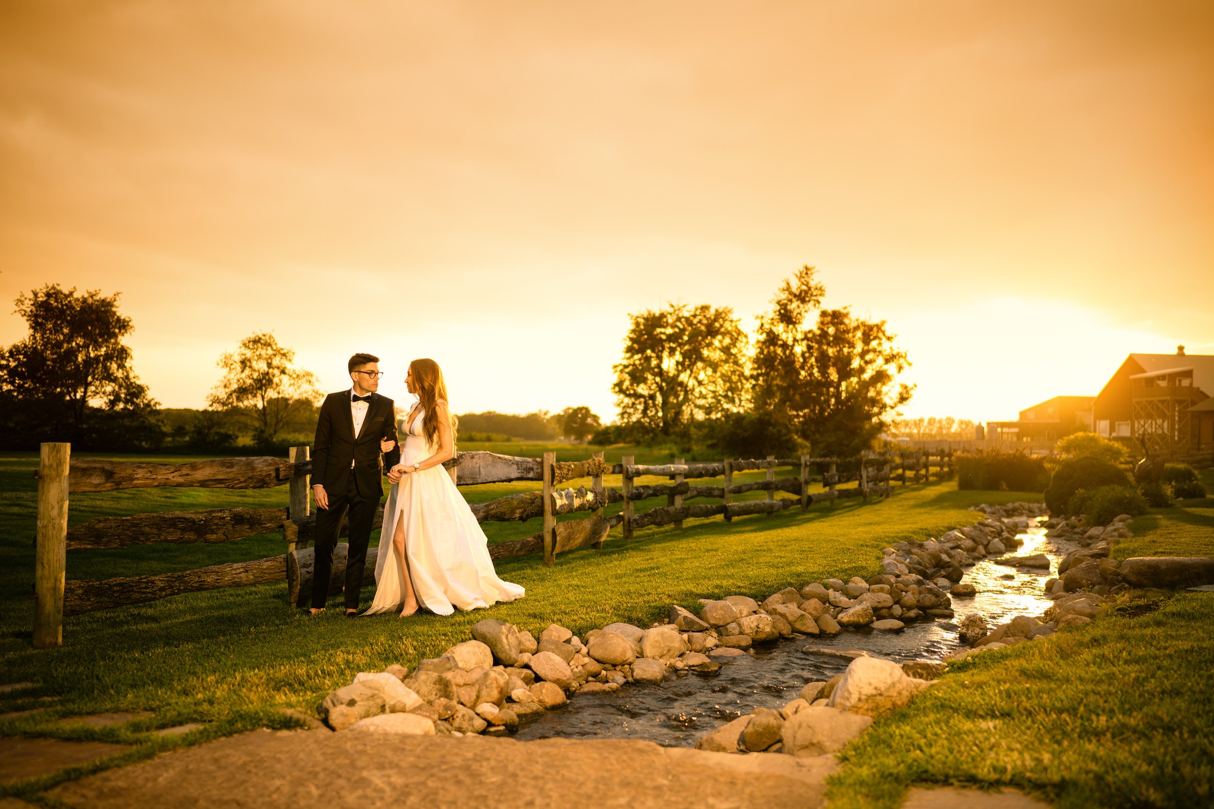 A couple dressed in formal wedding attire walks together beside a wooden fence and rocky stream at sunset, captured by a Northwest Indiana and Chicago wedding photographer in a beautiful rural outdoor setting.