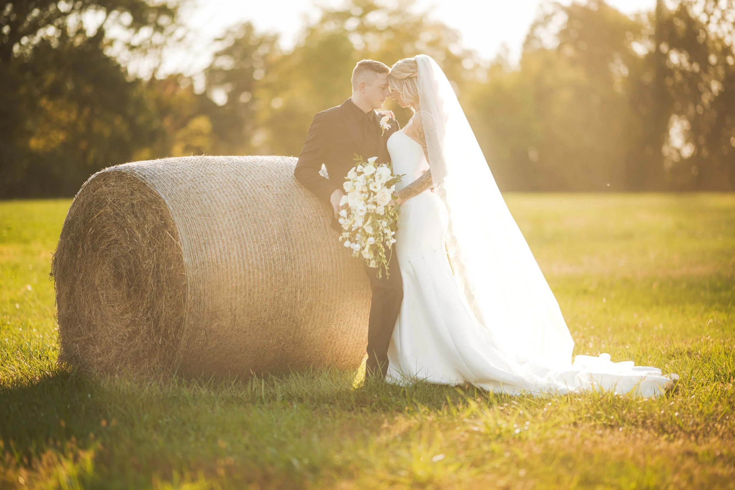 A bride and groom stand together in a grassy field next to a large hay bale, with sunlight streaming through trees in the background—captured beautifully by a Northwest Indiana and Chicago wedding photographer.