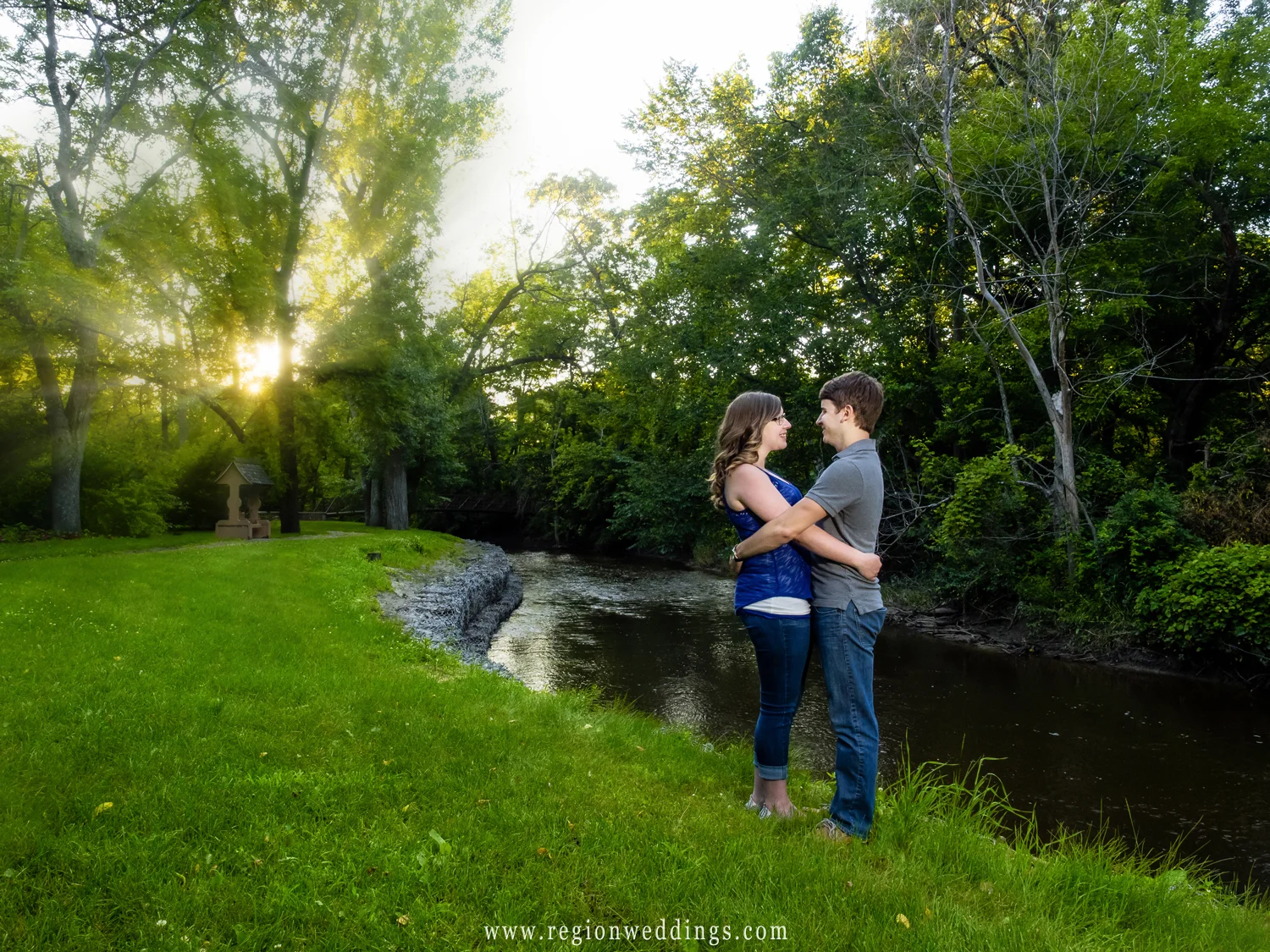 The sun shines through the trees as a young couple embraces along the river.