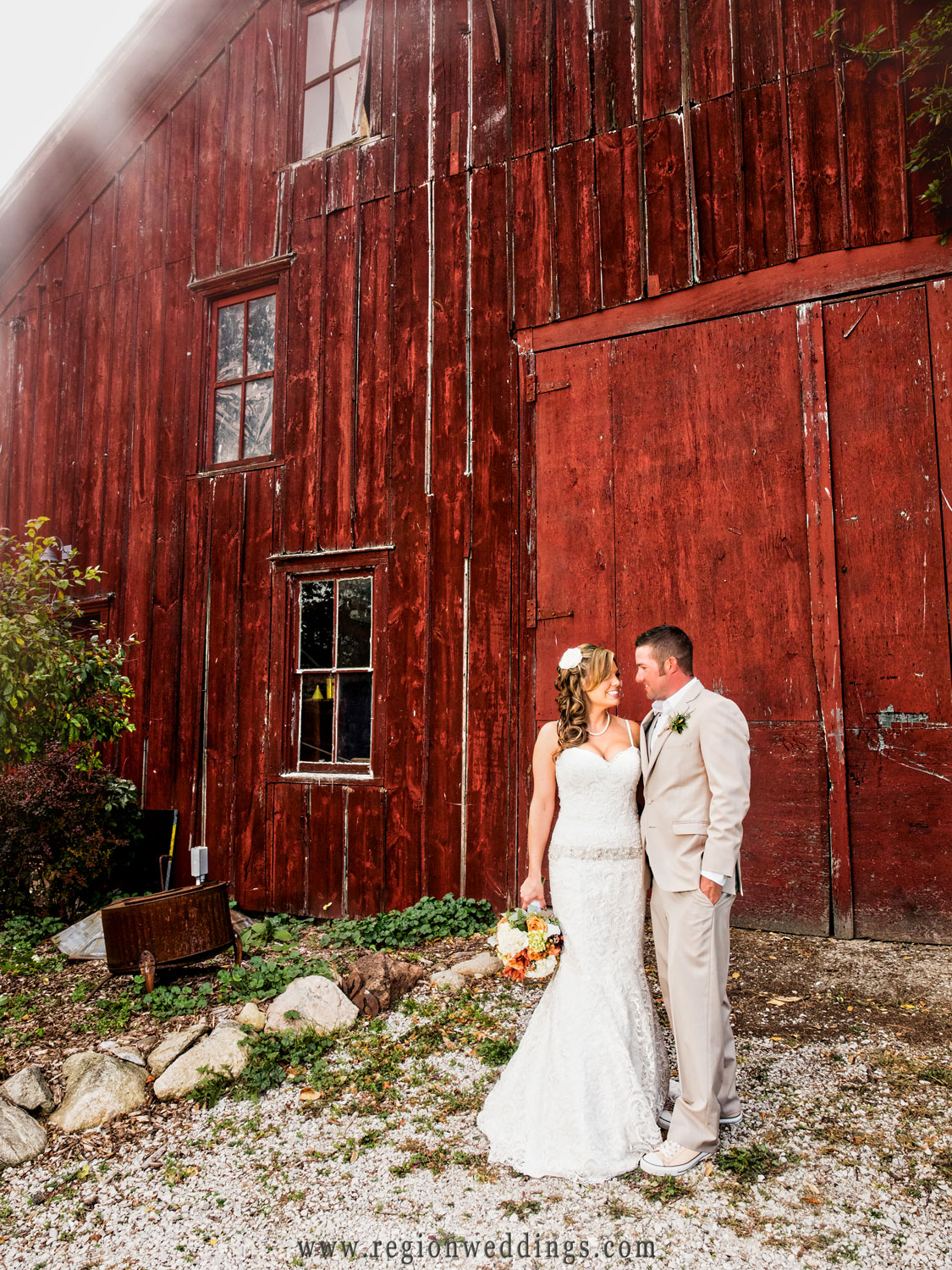 Sun rays illuminate the behind the red barn as the bride and groom gaze at each other.