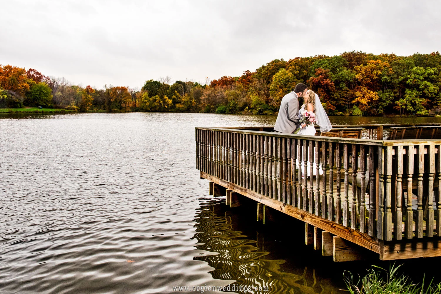 Fall colors pop in the background as the bride and groom kiss on the pier.