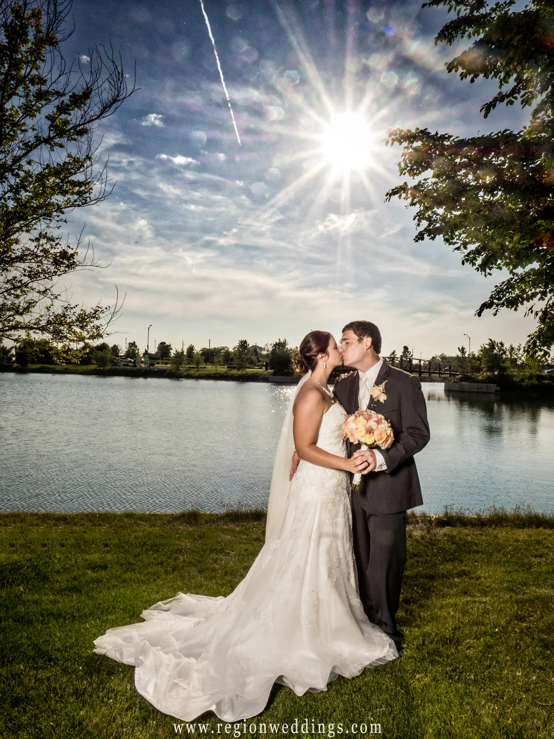 The bride and groom kiss underneath a sunburst after their outdoor summer wedding.