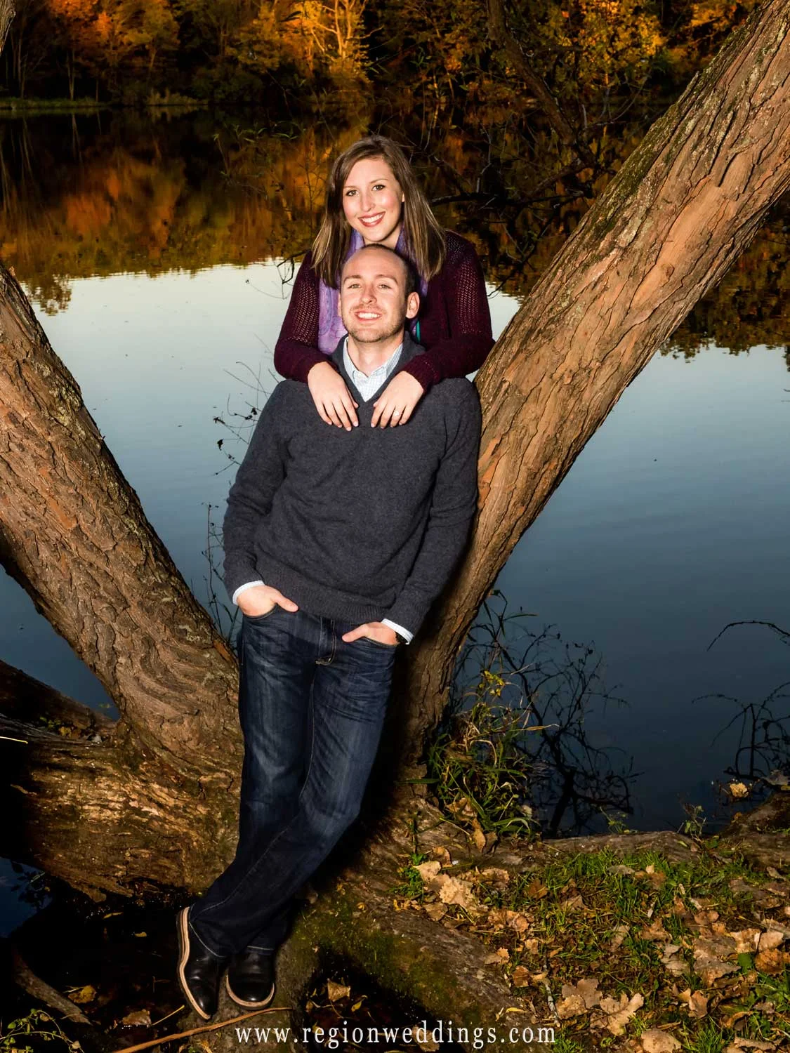 A portrait of an engaged couple perched alongside an old tree with Lemon Lake behind them.