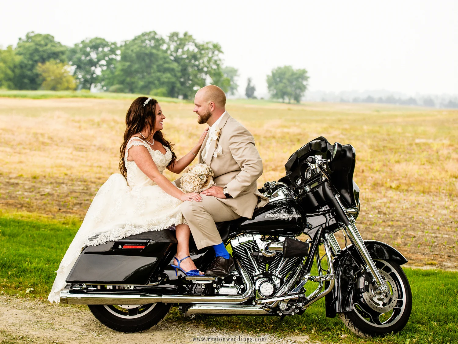 The bride and groom face each other on their Harley Davidson bike with farmland as their backdrop.