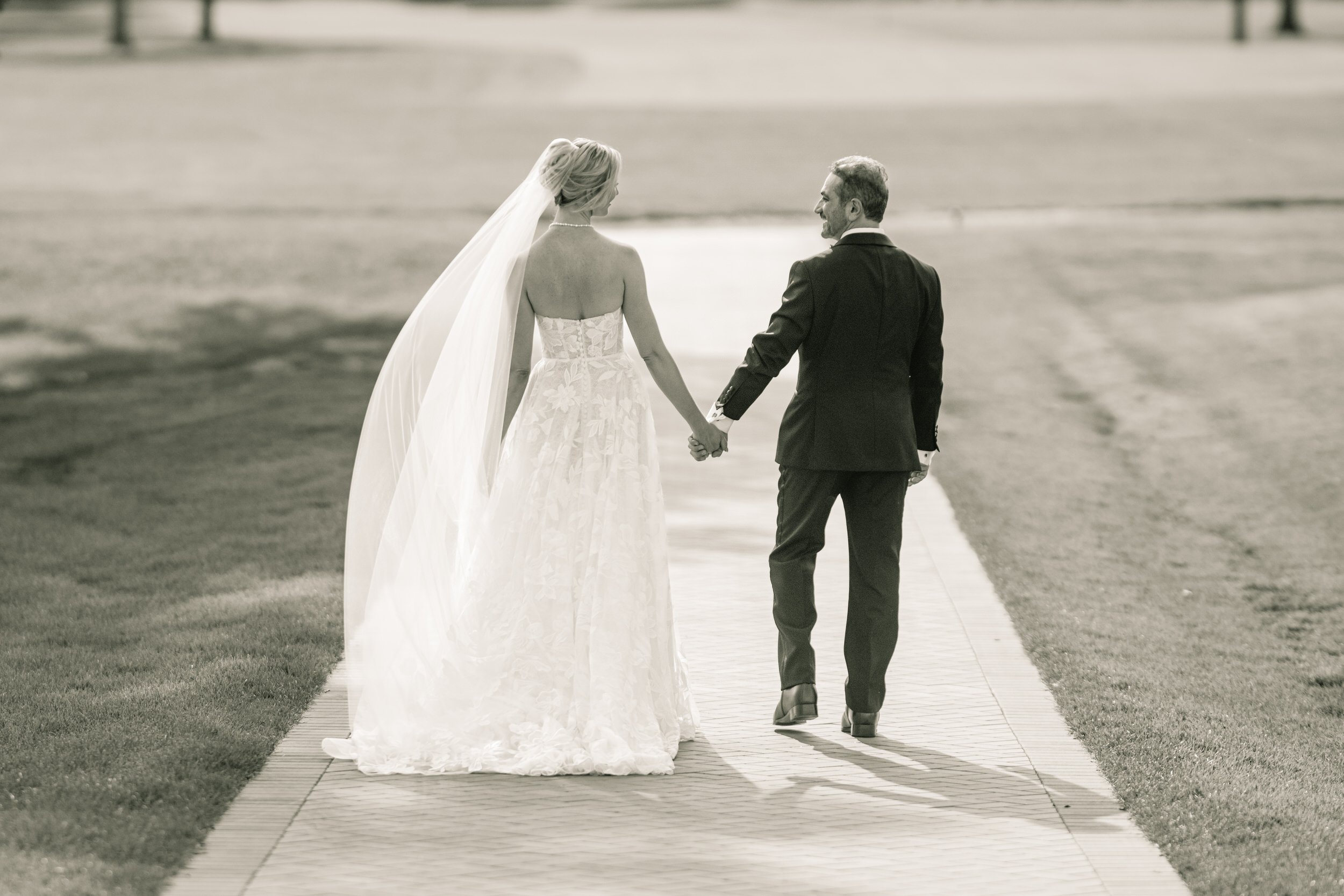 A black and white wedding photo of a bride and groom walking away from camera along a pathway at Midlothian Country Club golf course.