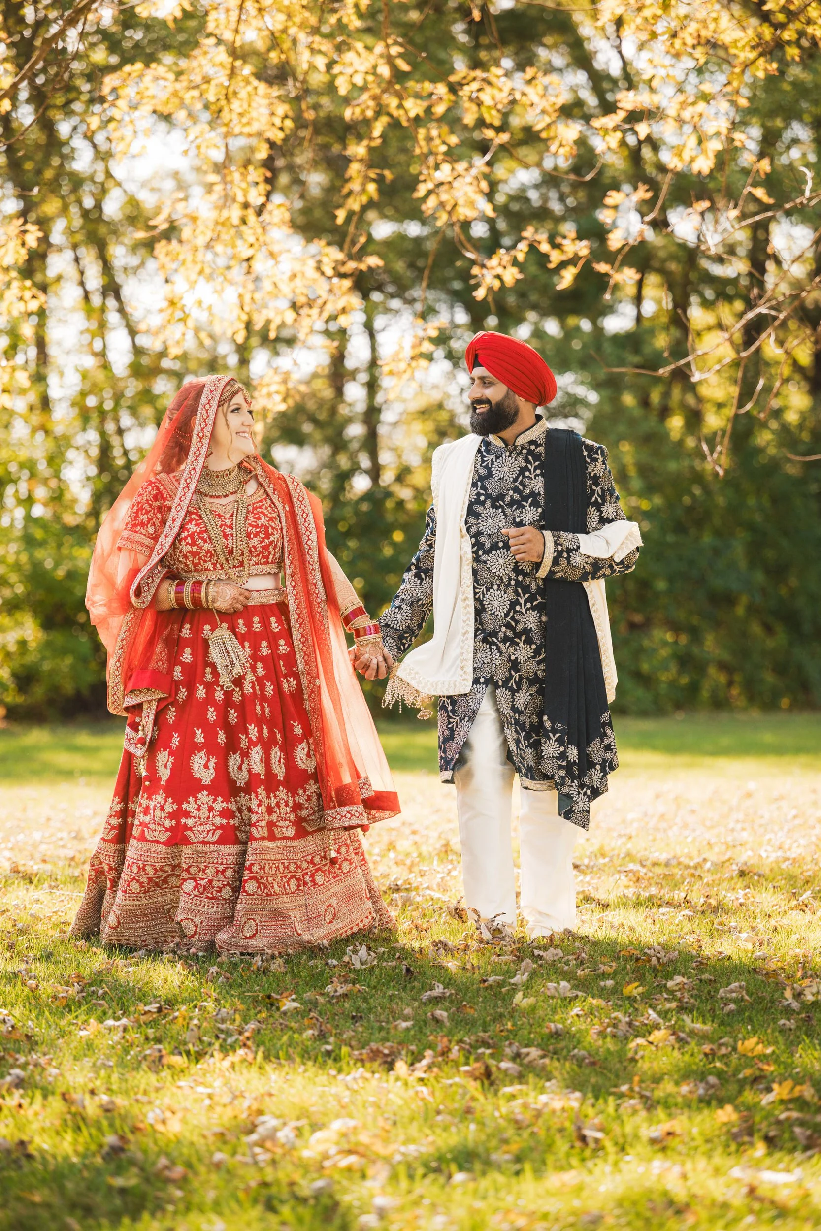 A couple in traditional Indian attire stands outdoors, holding hands and smiling at each other in a sunlit, grassy area with trees—captured by a Northwest Indiana and Chicago wedding photographer.