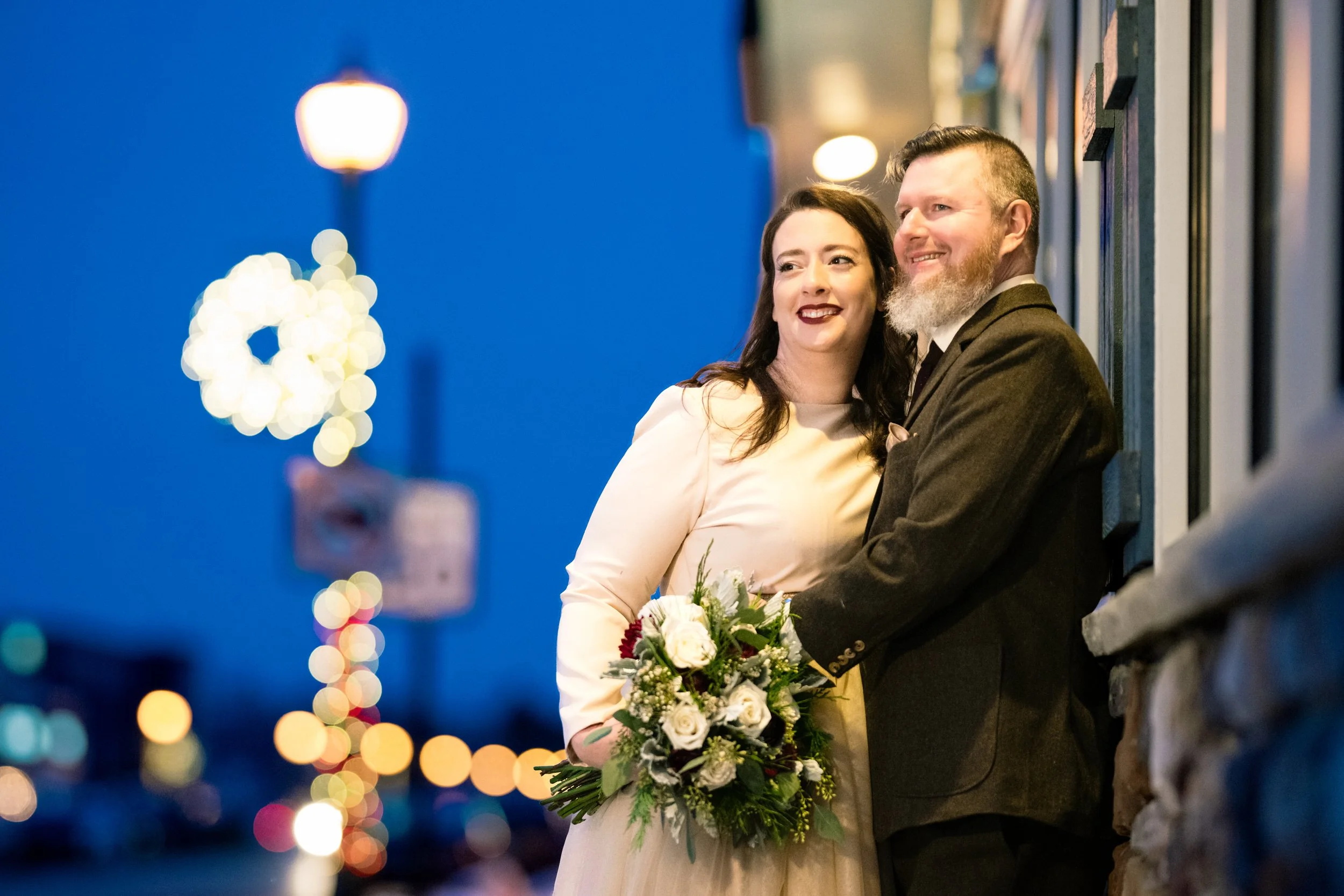 A couple in wedding attire stands outside at dusk, smiling and holding a bouquet, with blurred city lights in the background—captured beautifully by a Chicago wedding photographer serving Northwest Indiana.