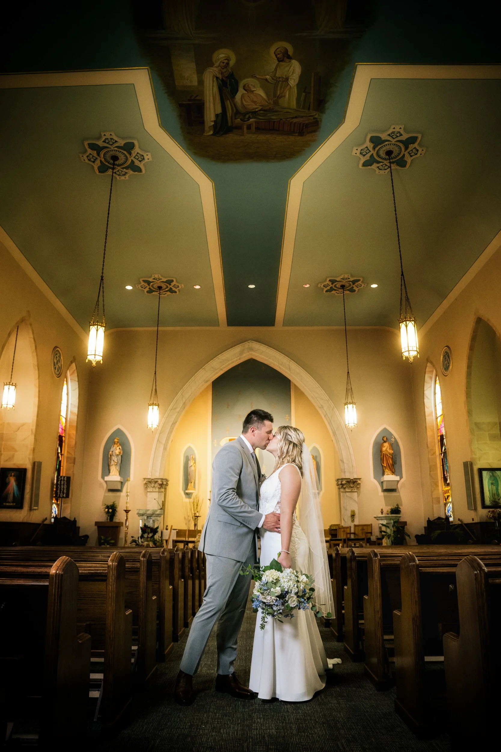 A bride and groom stand facing each other in the aisle of a church, holding hands and a bouquet, with decorative lighting and religious artwork overhead—captured by a Northwest Indiana and Chicago wedding photographer.