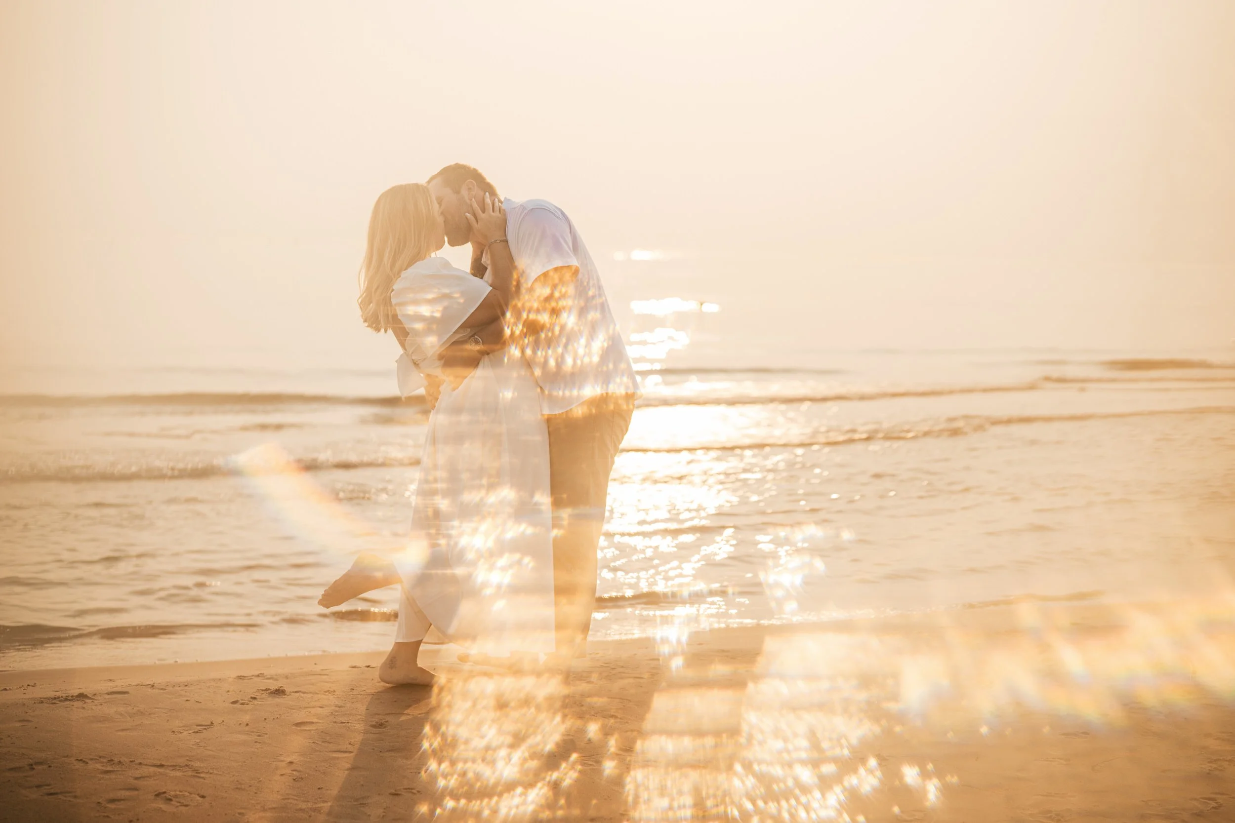 A couple stands on the sandy shores of Silver Lake Beach at golden hour, embracing and kissing near the shoreline as sunlight reflects off the water in southwest Michigan.
