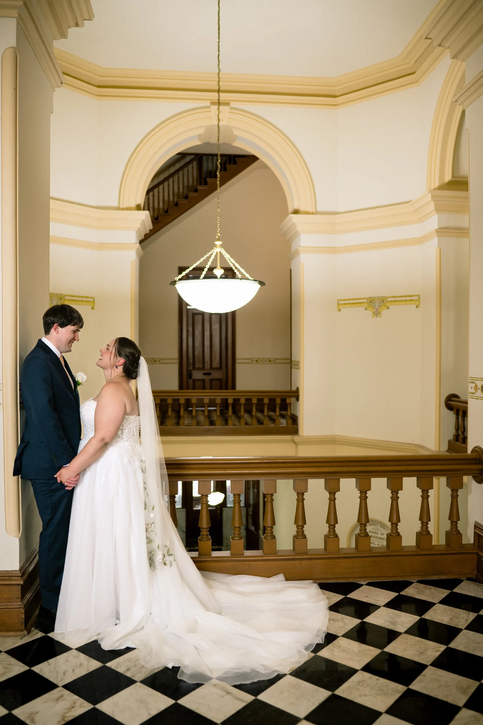A bride and groom stand holding hands in an elegant hallway with checkered floors, wooden railings, and an arched architectural background, beautifully captured by a Northwest Indiana and Chicago wedding photographer.