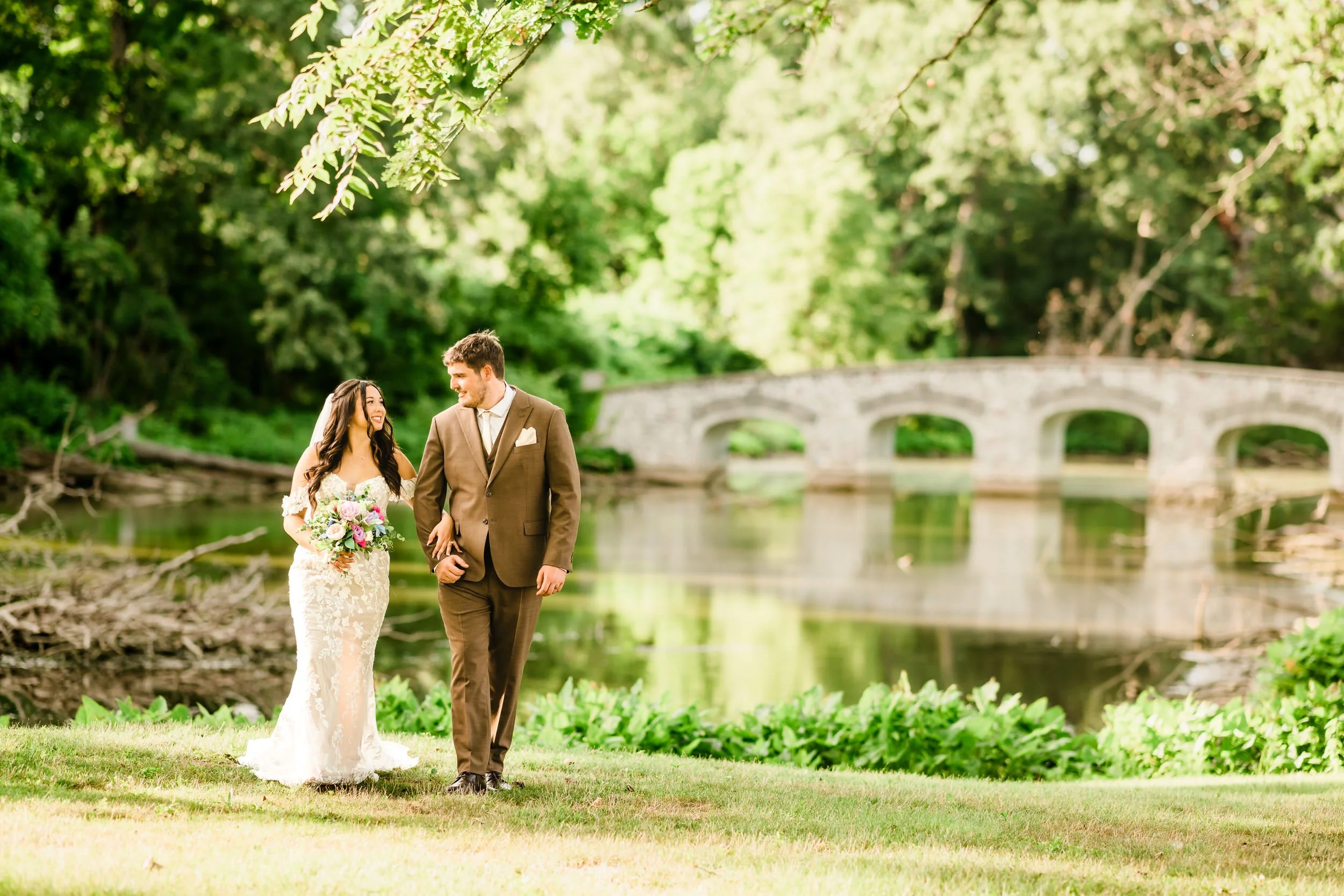 A bride and groom walk hand in hand on grass near a pond, with a stone bridge and trees in the background, beautifully captured by a Northwest Indiana and Chicago wedding photographer.
