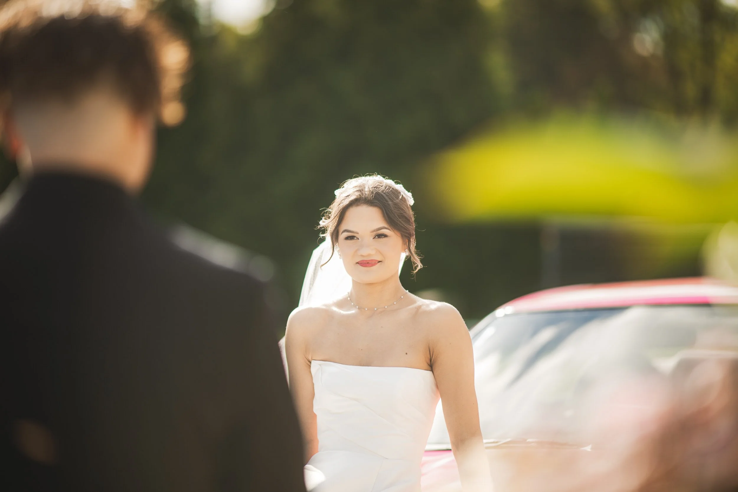 A bride in a white dress stands outdoors, facing a person in a dark suit, with greenery and a red car in the background—captured by a Northwest Indiana and Chicago wedding photographer.