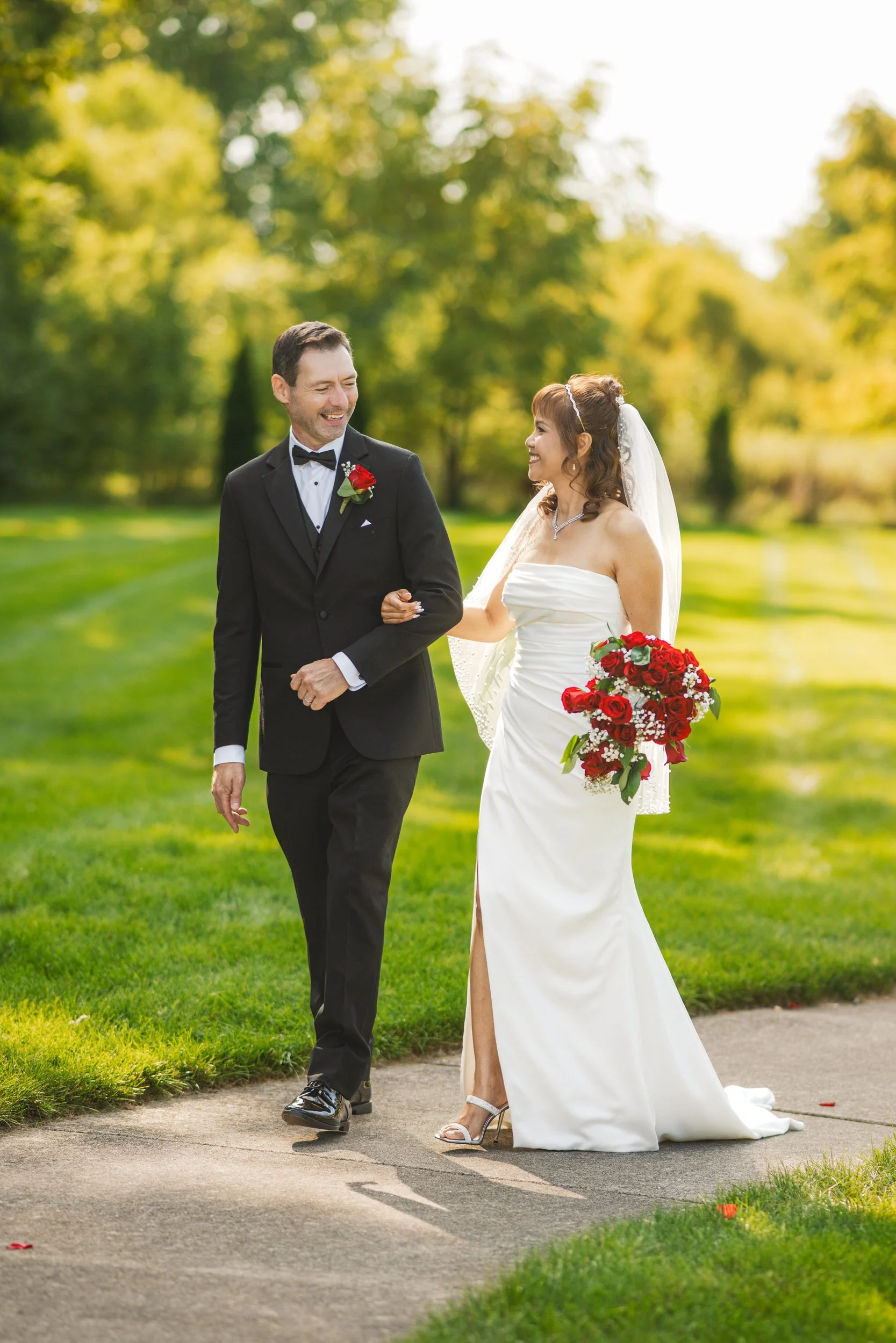 A bride in a white dress and groom in a black suit walk arm-in-arm outdoors on a sunny day, holding a red and white floral bouquet, beautifully captured by a Northwest Indiana and Chicago wedding photographer.