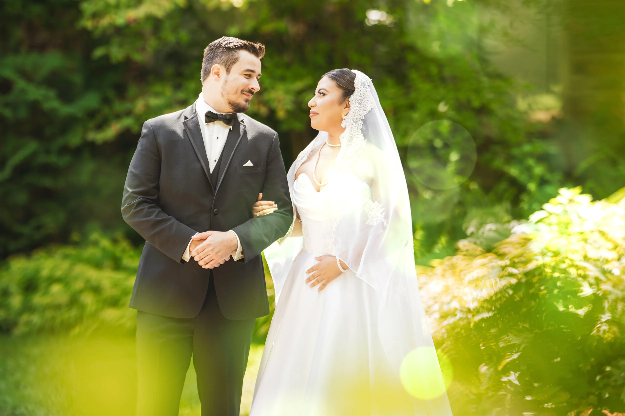 A bride in a white dress and veil stands arm in arm with a groom in a black tuxedo outdoors, surrounded by greenery and sunlight—captured beautifully by a northwest Indiana and Chicago wedding photographer.