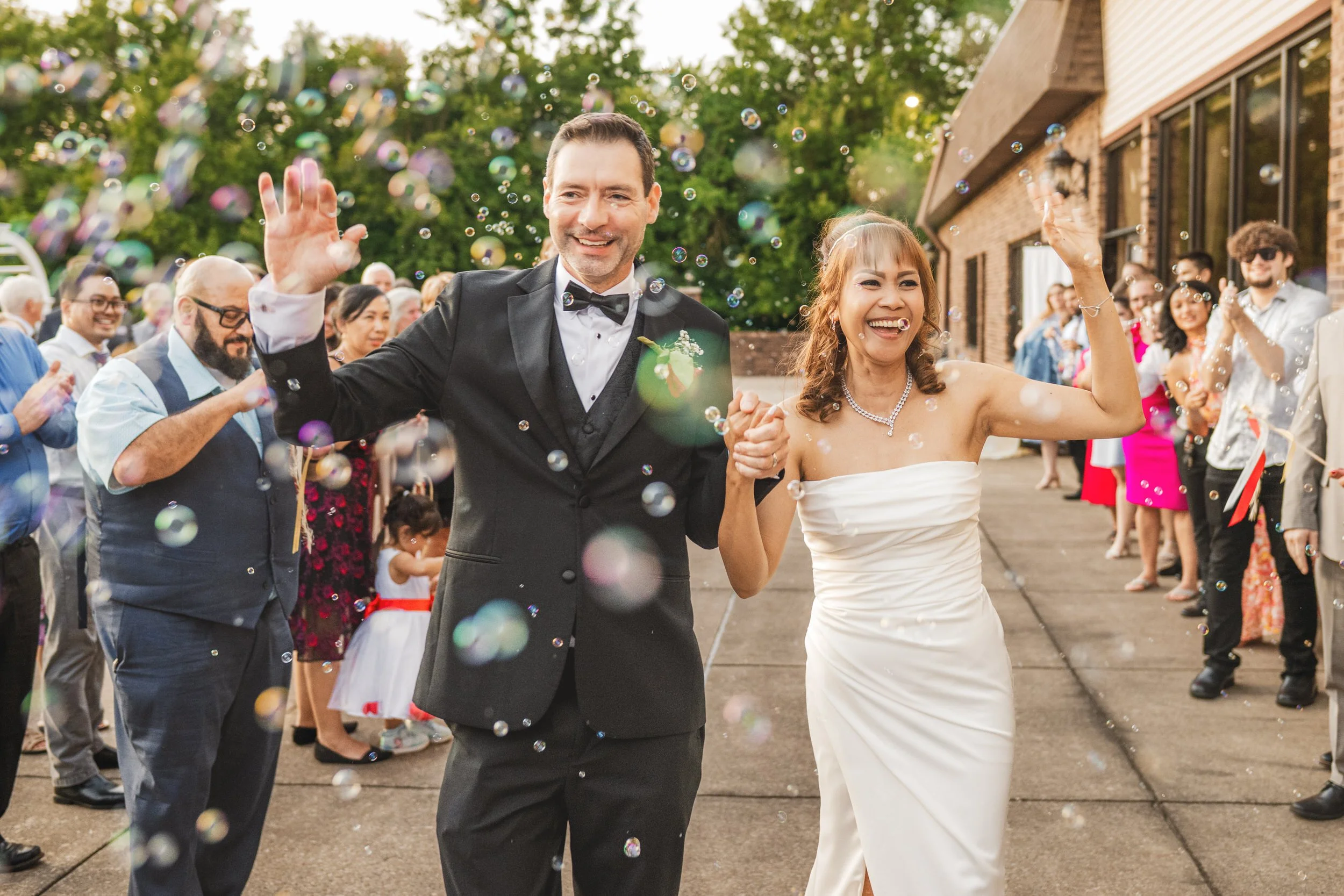 A bride and groom in formal attire walk outside holding hands and smiling while guests blow bubbles around them, captured beautifully by a northwest Indiana and Chicago wedding photographer at a joyful celebration.