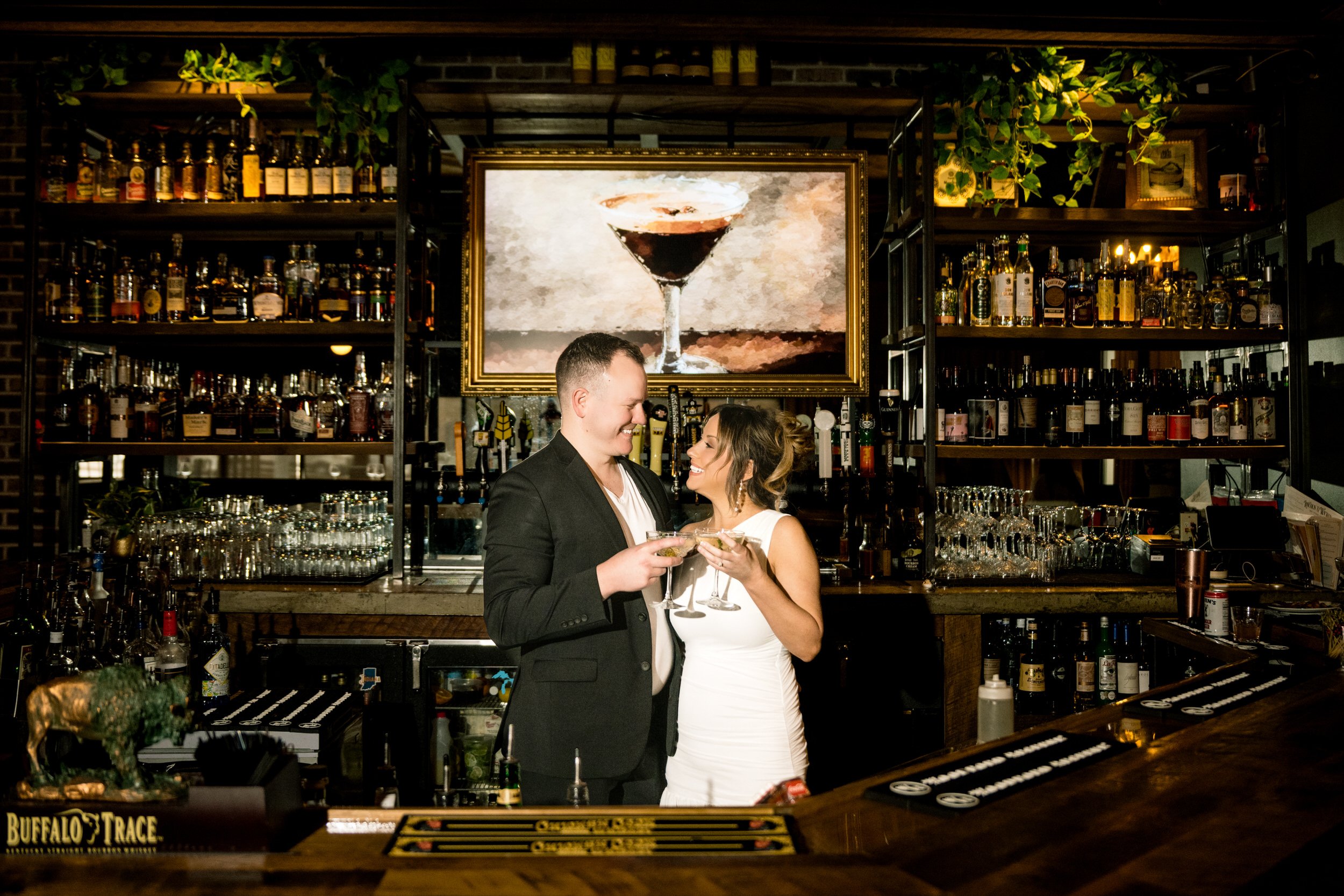 A romantic engagement photo of a couple enjoying a martini behind the bar of a classy restaurant.