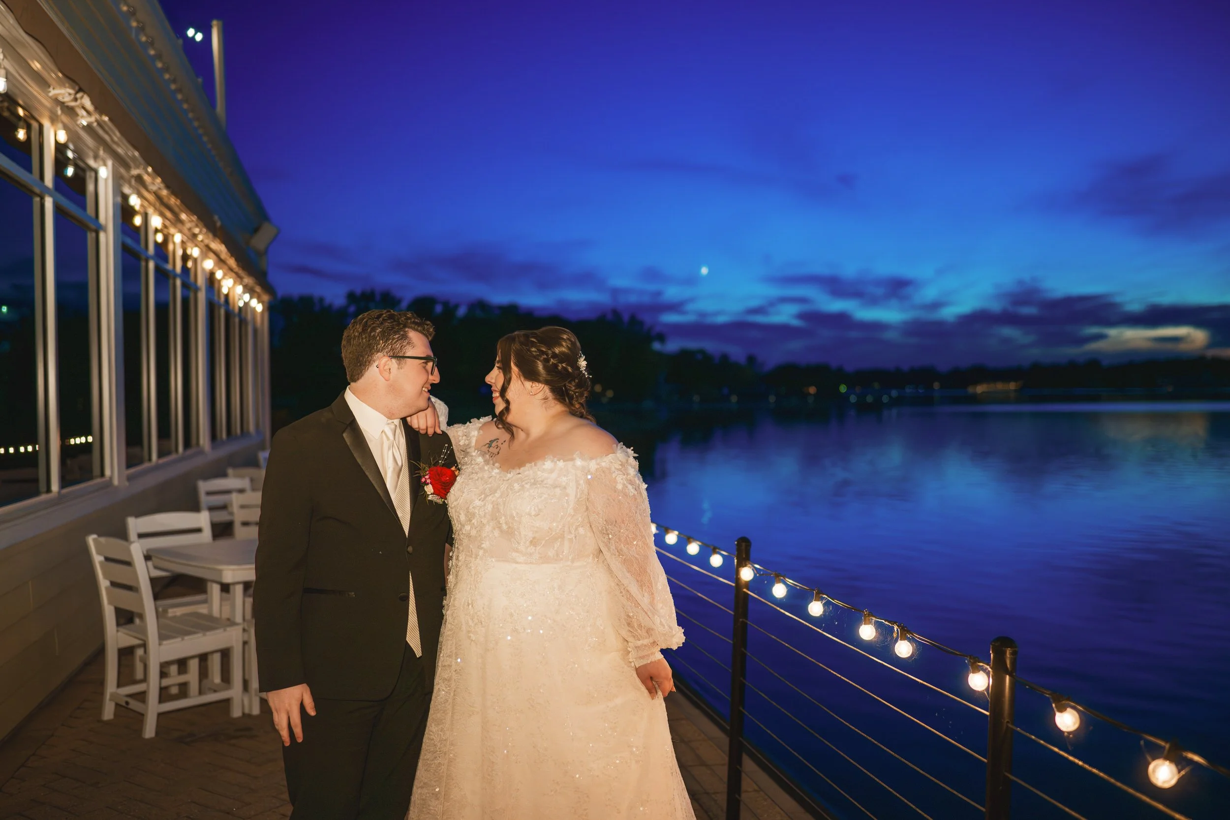 A couple dressed in formal wedding attire stand on a lakeside deck at dusk, looking at each other with string lights and a deep blue sky in the background—a perfect moment captured by a Northwest Indiana and Chicago wedding photographer.