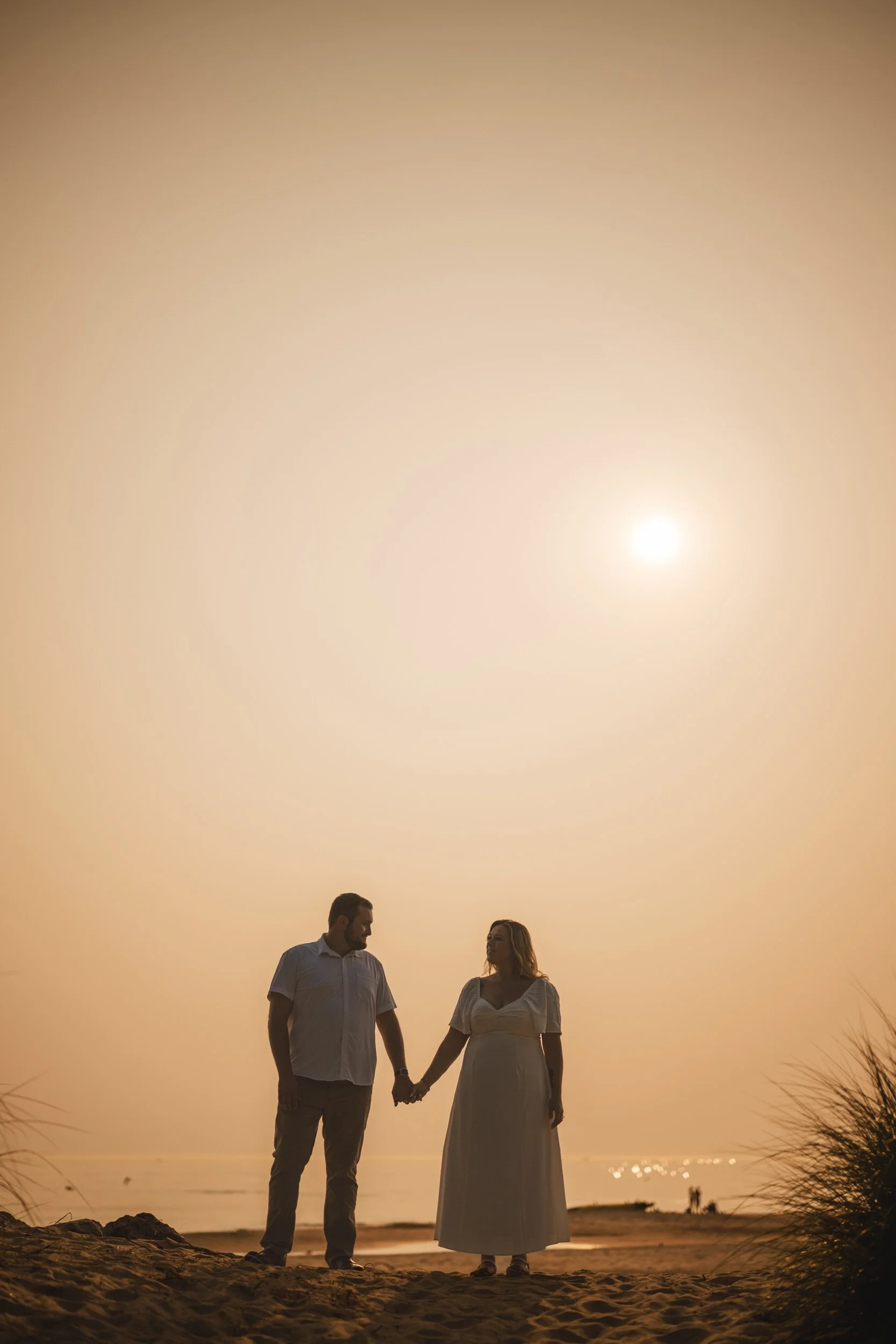 A couple stands on a sandy beach in southwest Michigan at sunset, holding hands and facing each other under a bright, golden hour sky.