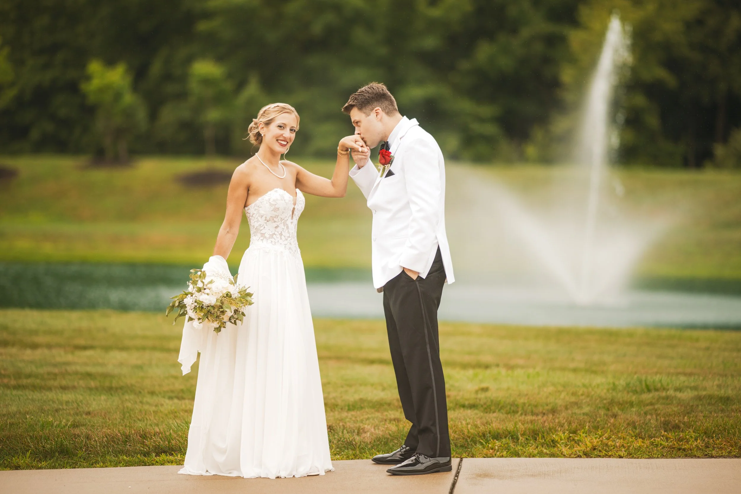 The groom kisses the bride's hand as she looks at the camera and a fountain of water spouts in the background.