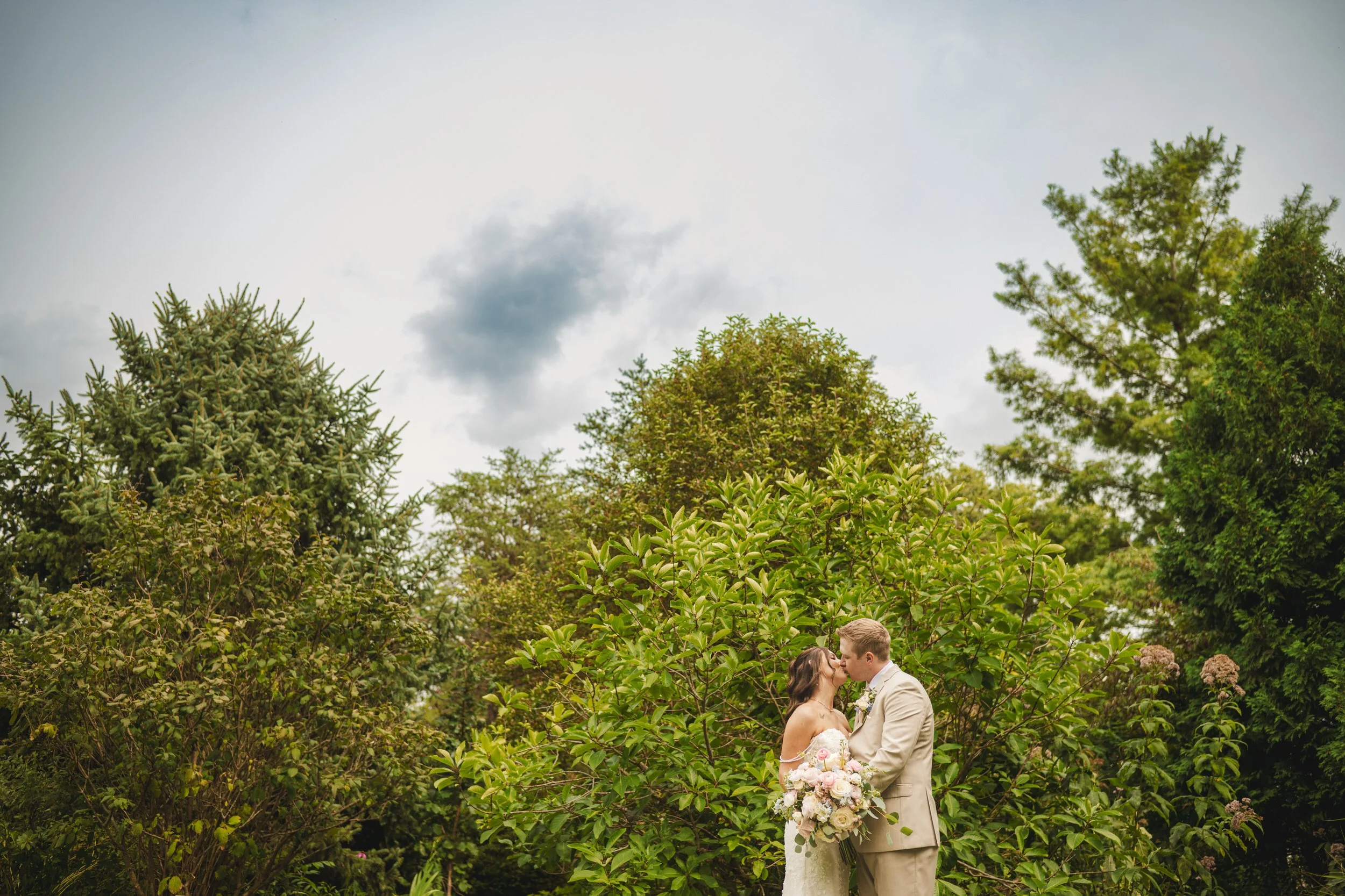 The bride and groom kiss with a puffy cloud above them as they stand surrounded by greenery.