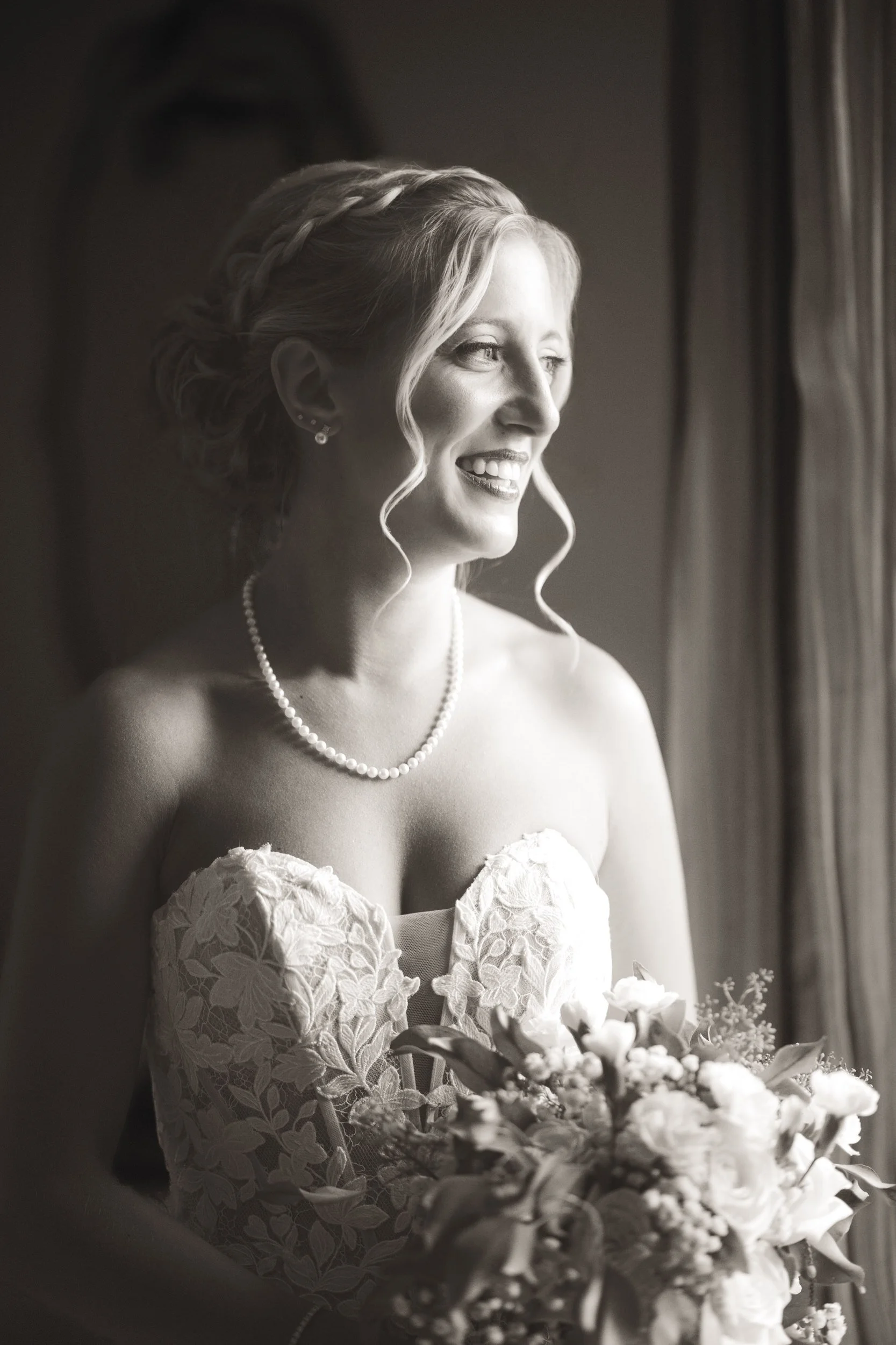 A black and white image of a bride holding flowers and looking out of the window at rain falling.