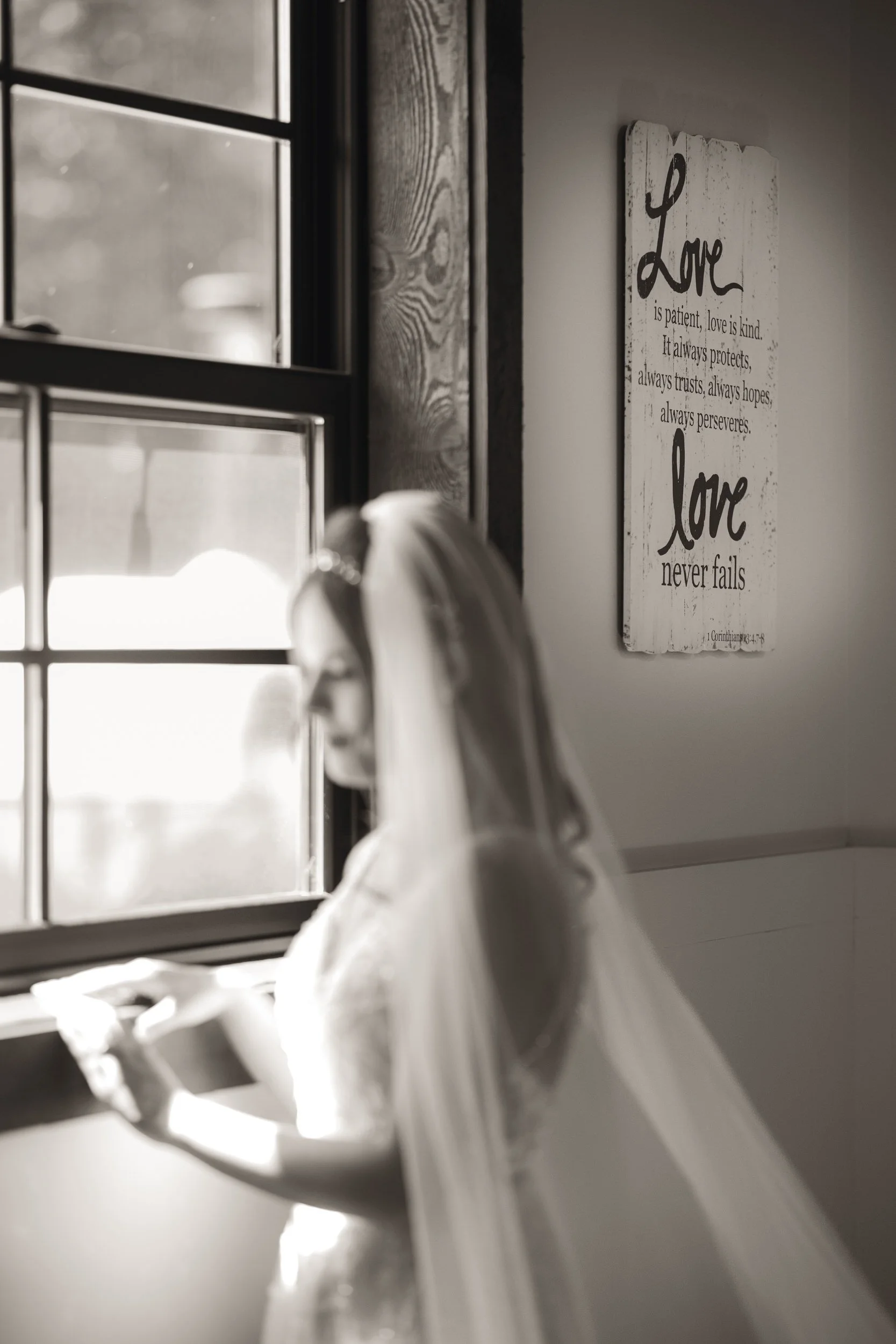 A bride in a veil stands by a window, reading a note; nearby, a wall sign displays "Love never fails." Captured by a northwest Indiana and Chicago wedding photographer.
