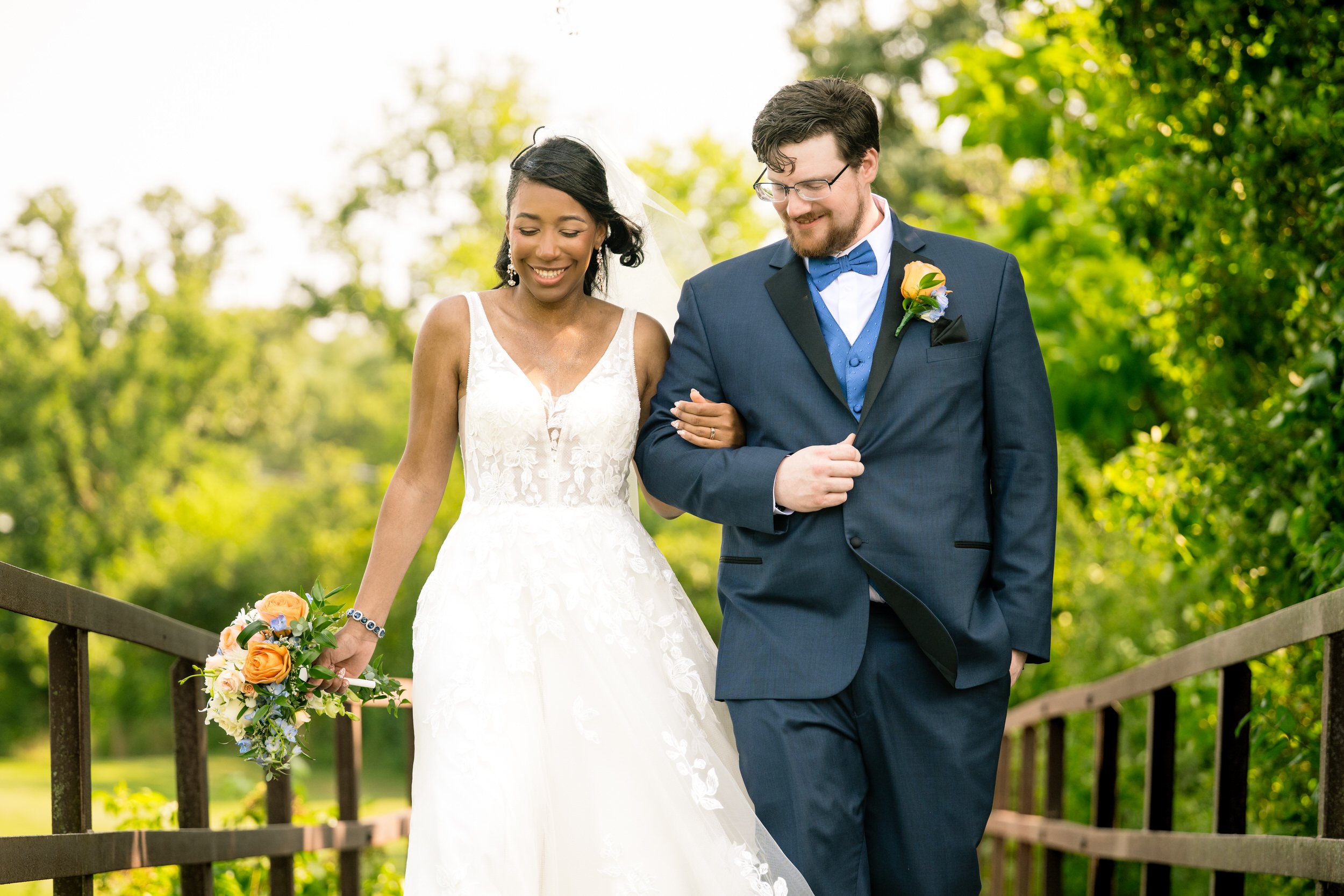 A bride and groom, dressed in formal wedding attire, walk arm in arm outdoors on a bridge surrounded by greenery. Captured by a northwest Indiana and Chicago wedding photographer, the bride holds a bouquet of orange and white flowers.