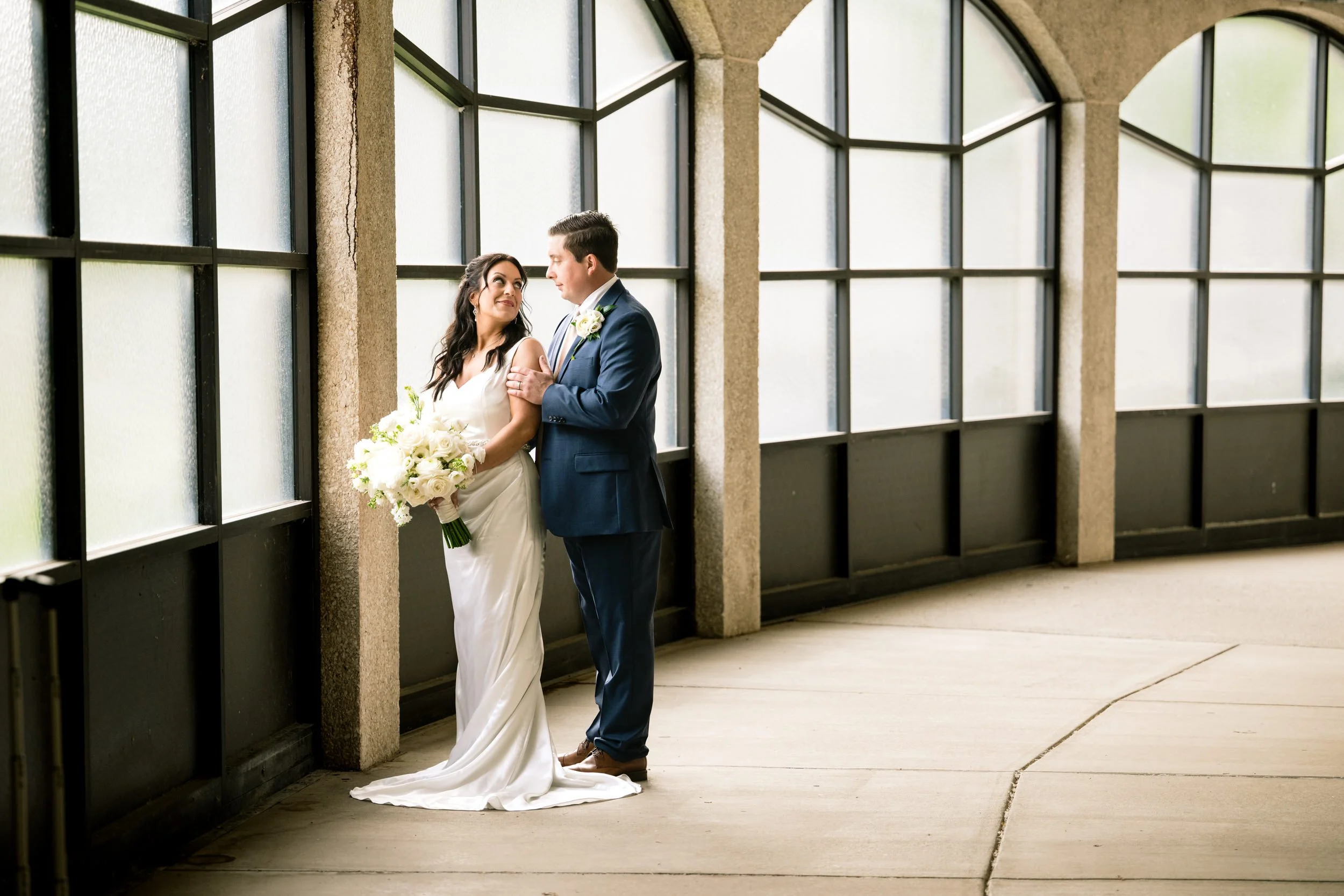 A bride in a white dress holding a bouquet stands next to a groom in a blue suit inside a hallway with large windows, captured beautifully by a Northwest Indiana and Chicago wedding photographer.