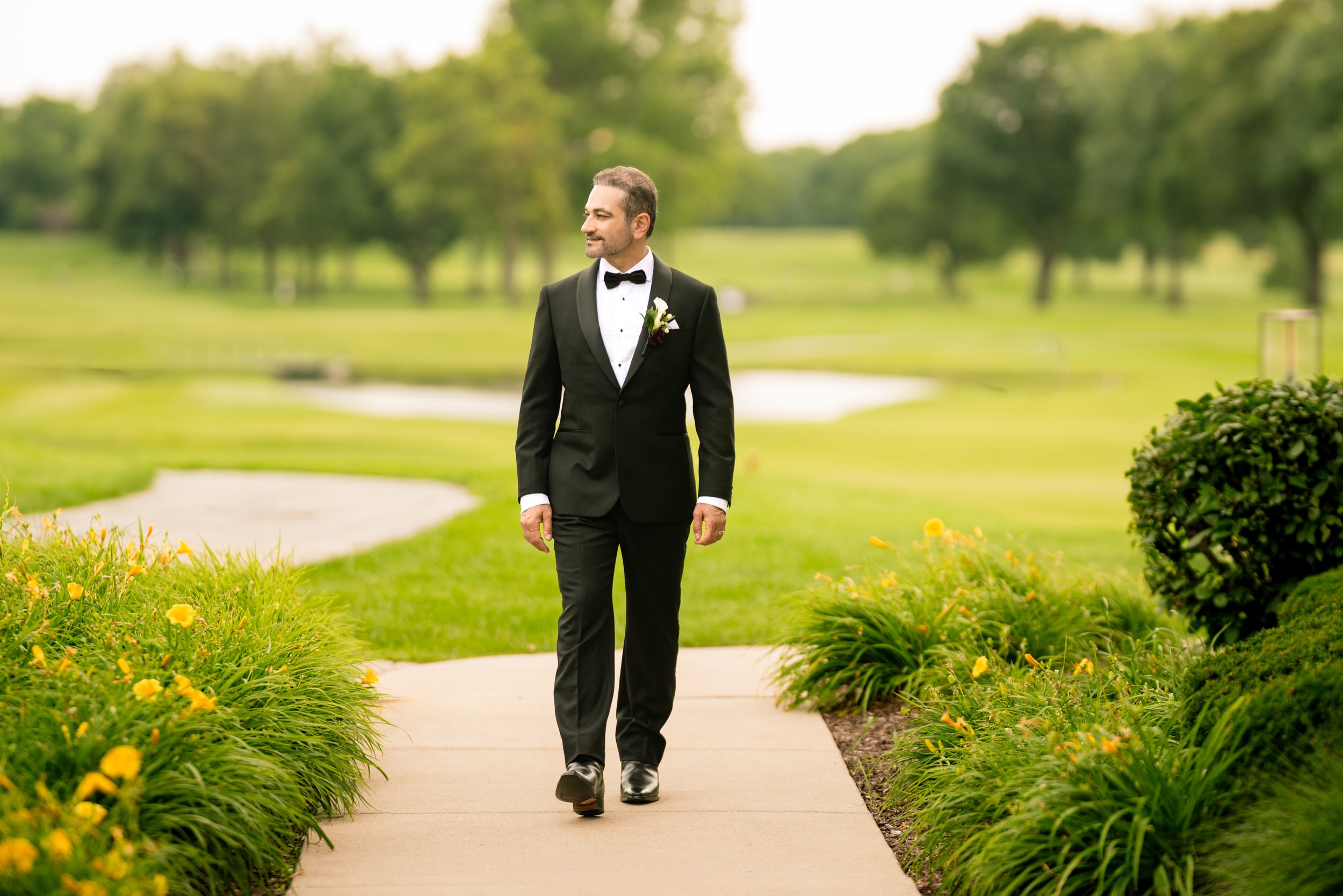 A dapper groom in a black suit walks with Midlothian Country Club golf course behind him.