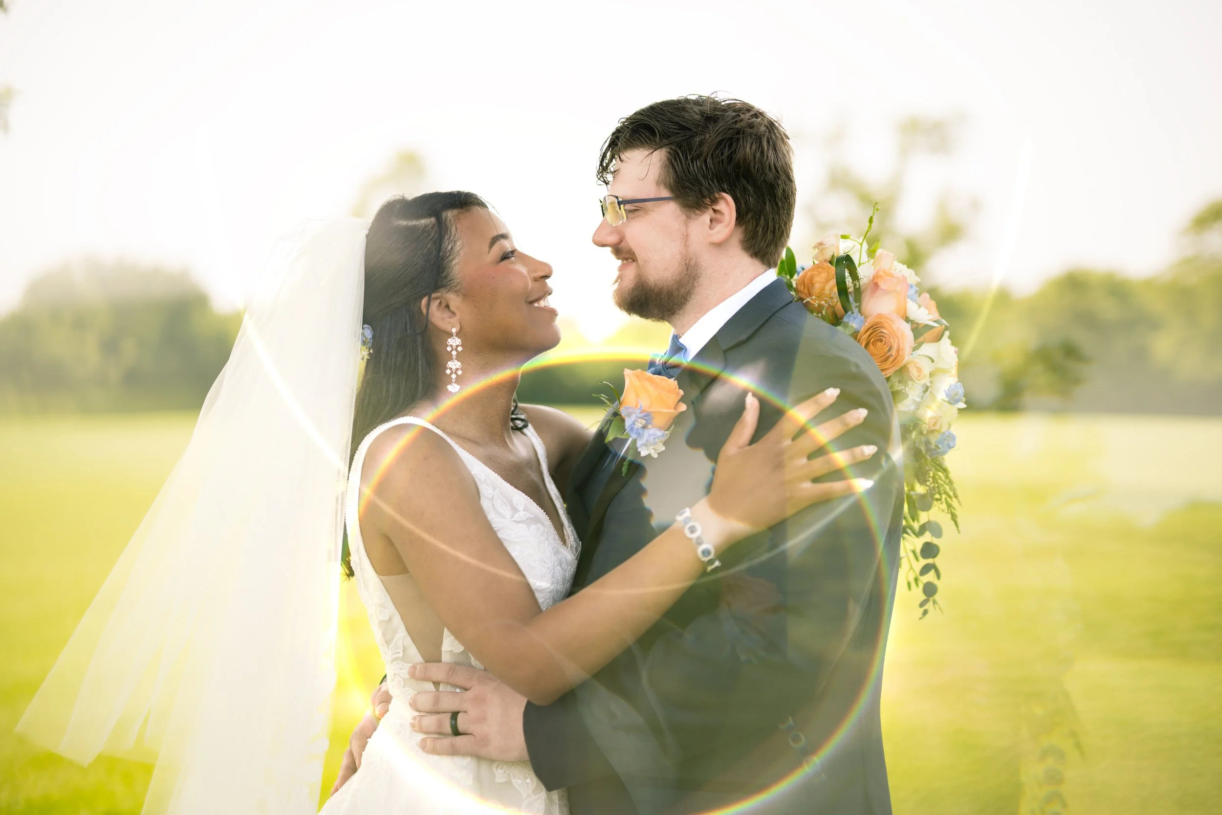 A bride and groom embrace outdoors, smiling at each other. The bride holds a bouquet and wears a veil. Captured by a northwest Indiana and Chicago wedding photographer, a circular light flare is visible over the image.