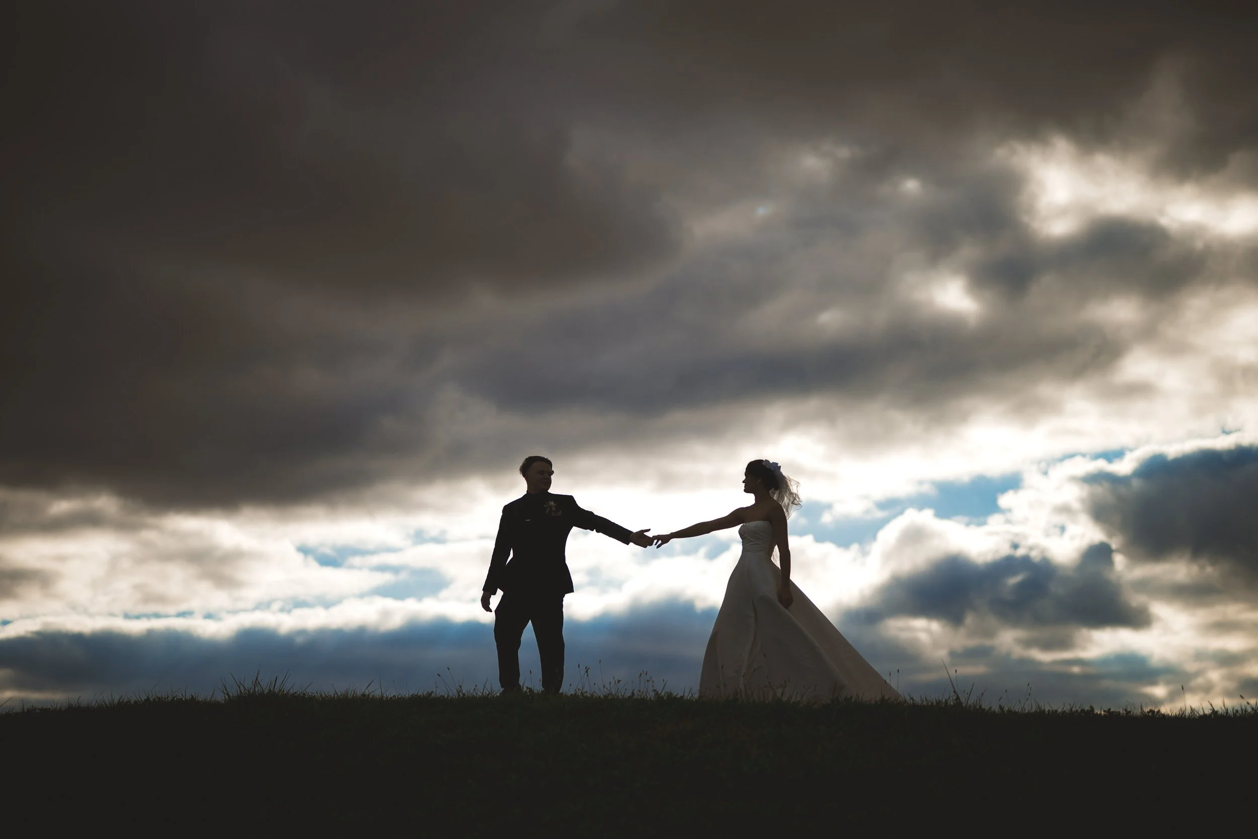 Silhouetted couple in wedding attire stand on a hill holding hands under a cloudy, dramatic sky with patches of sunlight, captured by a northwest Indiana and Chicago wedding photographer.