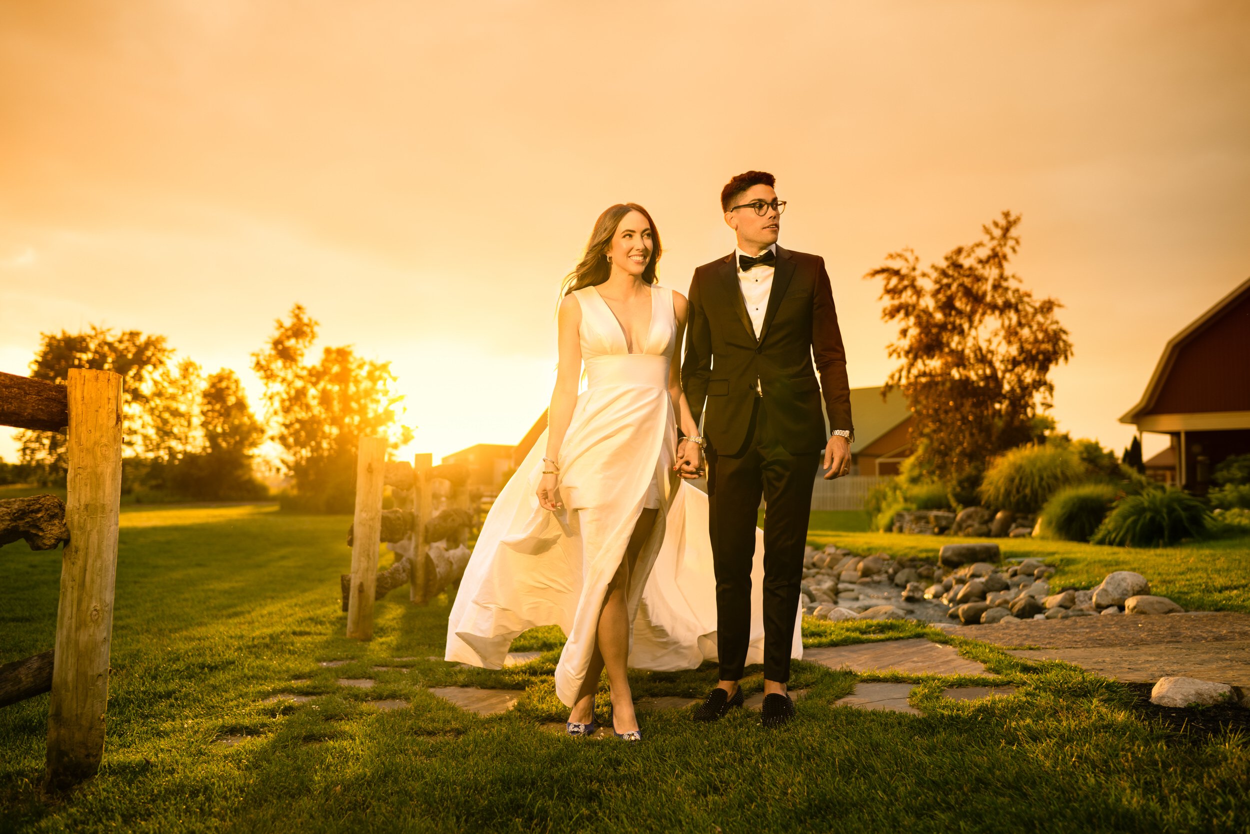 A couple dressed in formal attire walks hand in hand outdoors at sunset, with grass, rocks, and wooden fencing in the background—captured by a Northwest Indiana and Chicago wedding photographer.