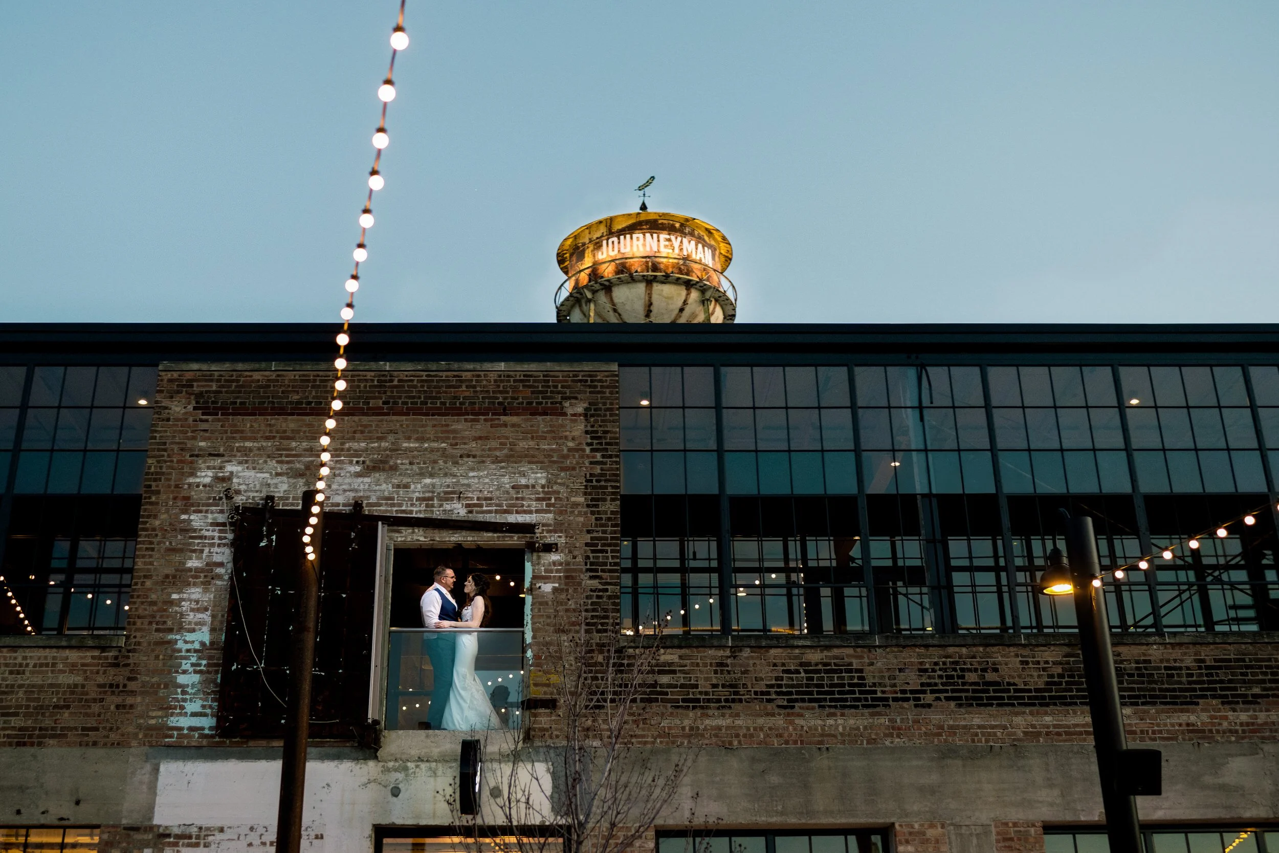 A couple stands on a small balcony of a brick industrial building at dusk, with string lights overhead and the "Journeyman" water tower in the background, beautifully captured by a Northwest Indiana and Chicago wedding photographer.