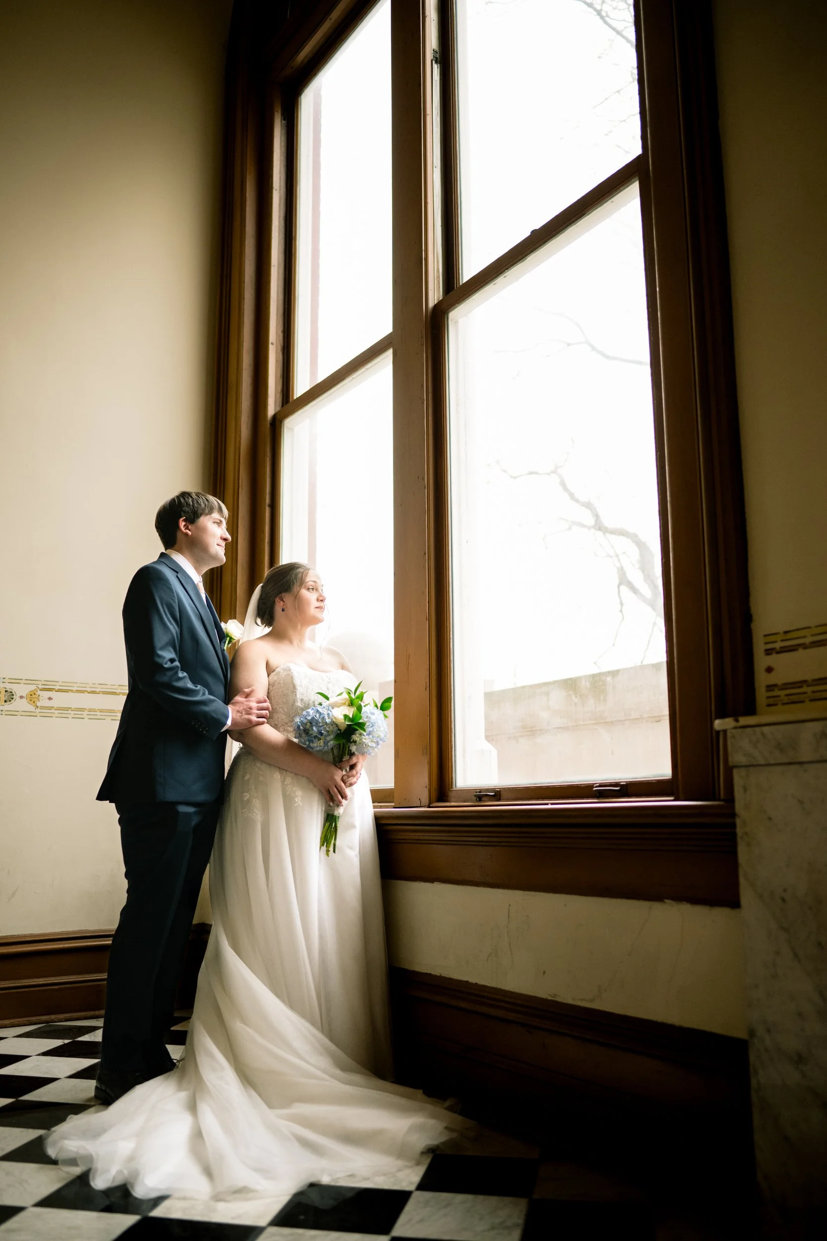 A bride and groom in formal attire stand together by a large window, looking outside. Captured by a Northwest Indiana and Chicago wedding photographer, the bride holds a bouquet, with checkered floors and tall ceilings completing the elegant scene.