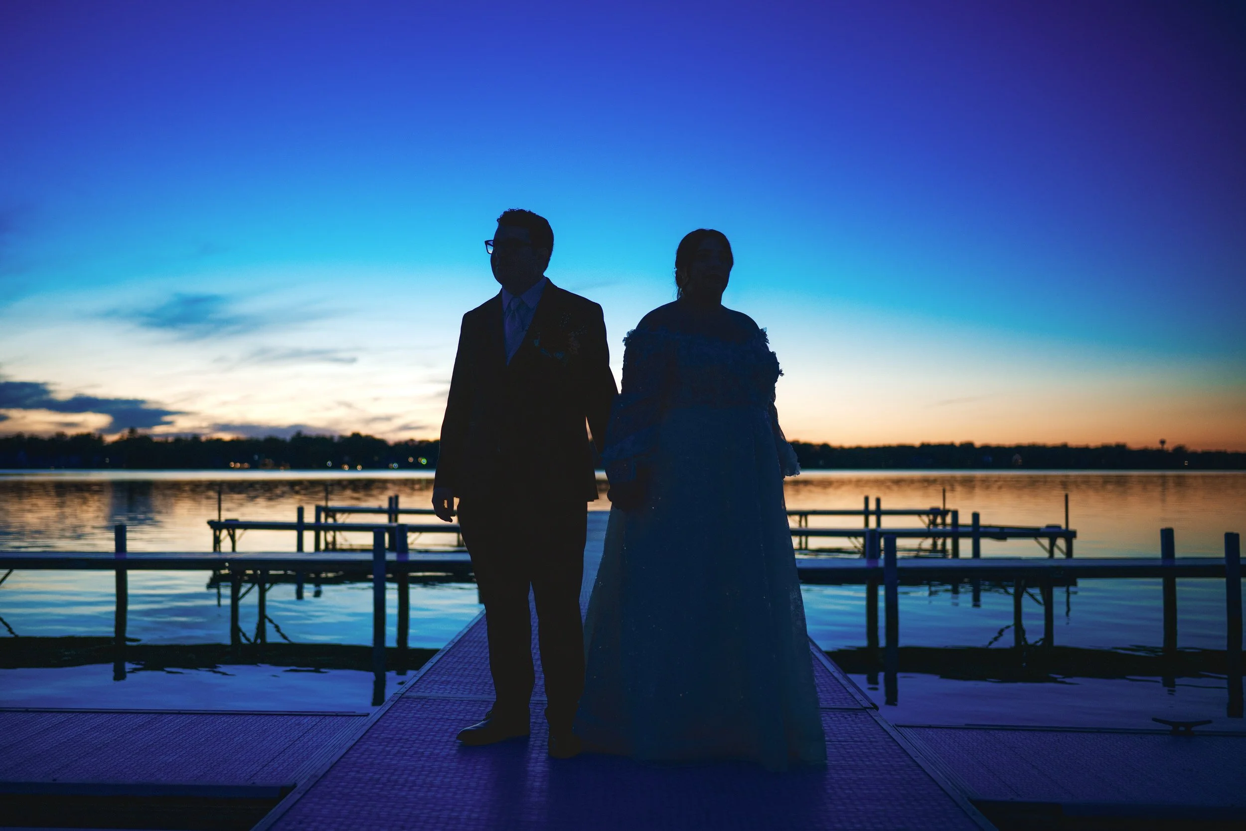Two people in formal attire stand hand in hand on a dock at sunset, silhouetted against a calm lake and a blue and purple sky—captured by a northwest Indiana and Chicago wedding photographer.
