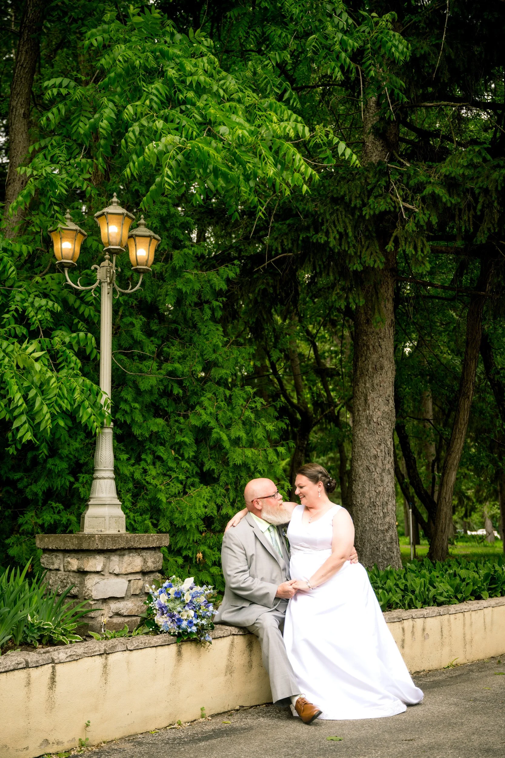 A bride in a white gown and a groom in a gray suit sit together on a stone ledge near a lamp post and trees, with a bouquet of flowers on the ground—captured by a Northwest Indiana and Chicago wedding photographer.