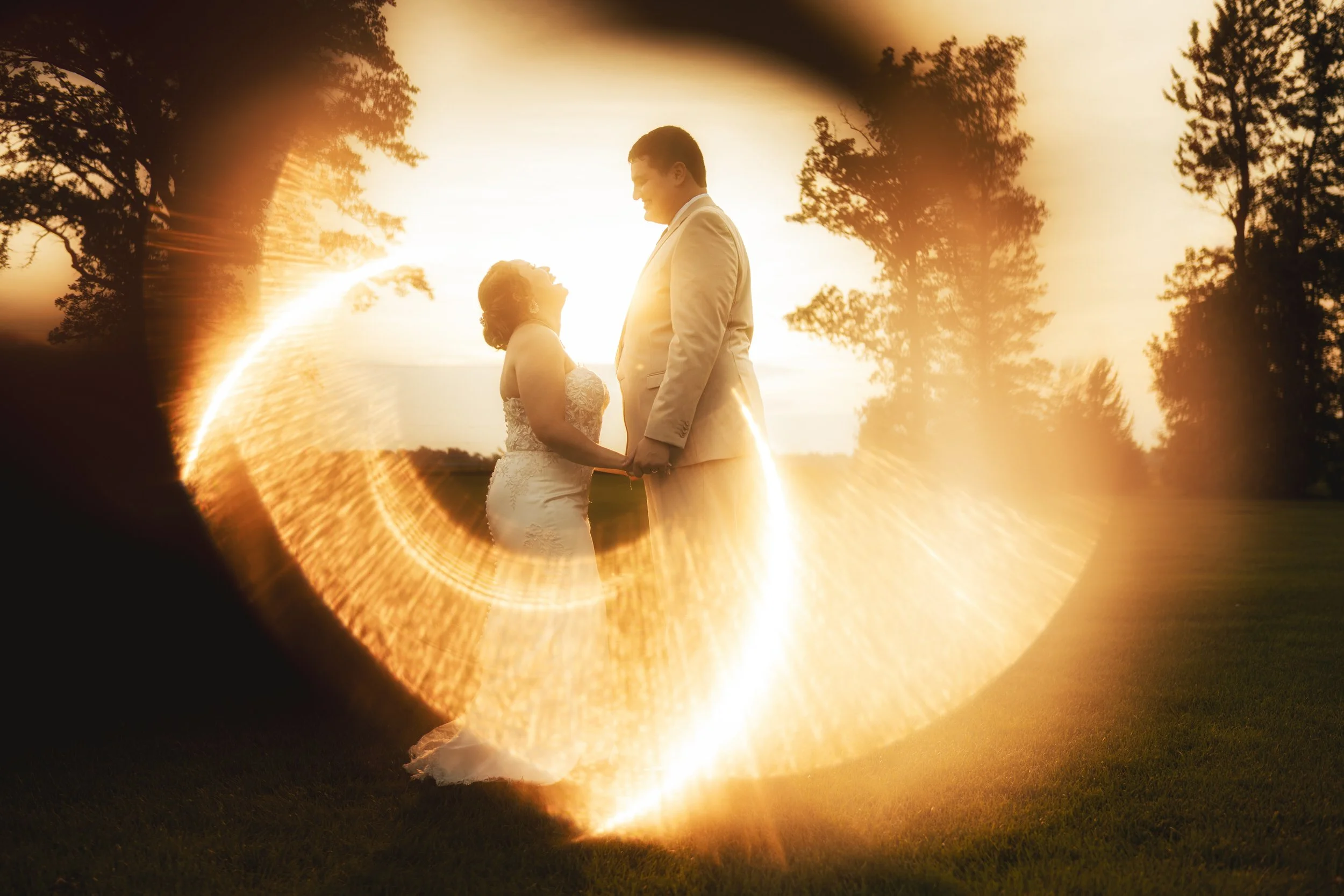 A bride and groom stand facing each other outdoors at sunset, surrounded by a circular golden light effect, with trees in the background—captured by a Northwest Indiana and Chicago wedding photographer.
