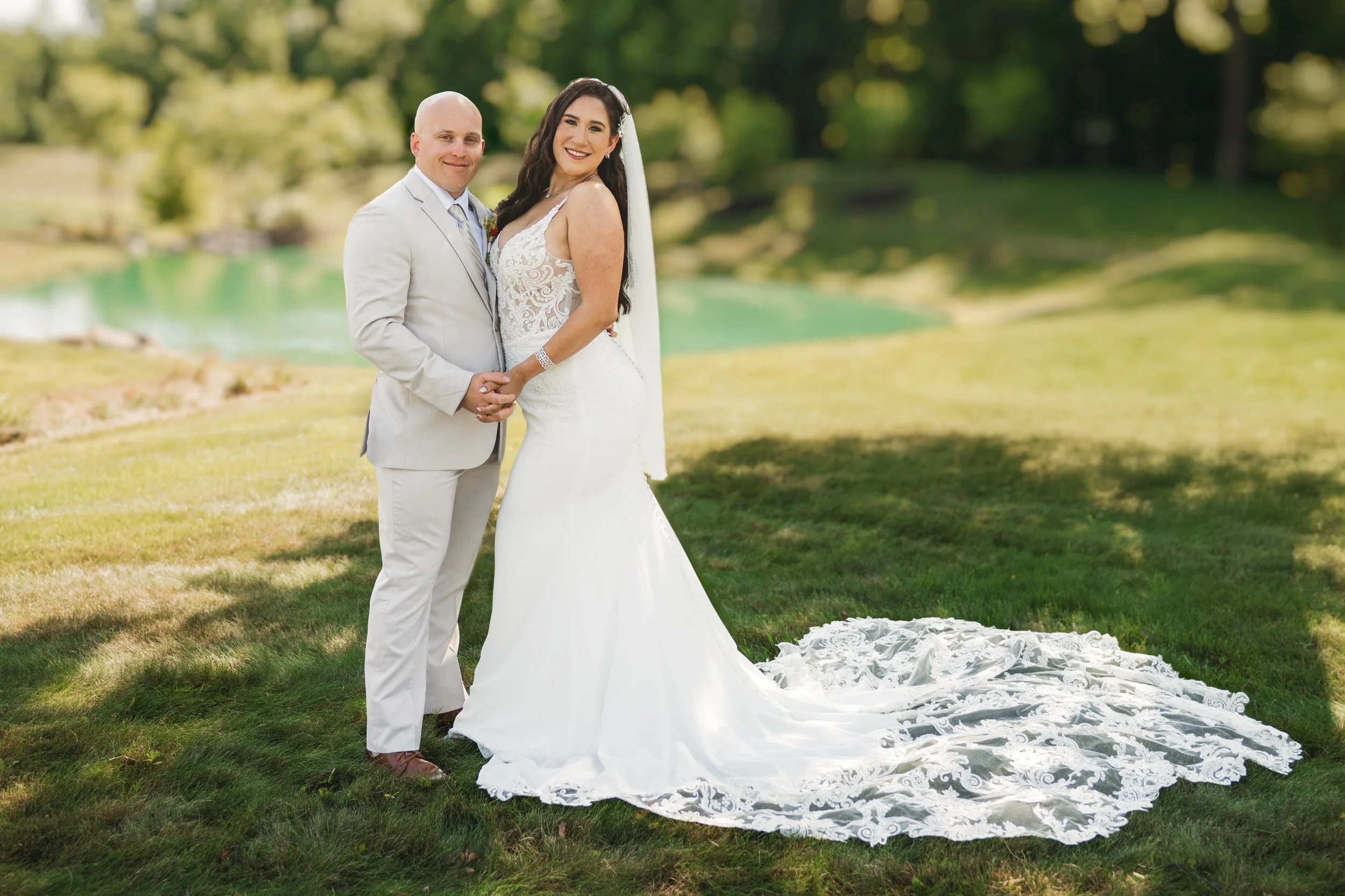 A bride in a white gown with a lace train and a groom in a light-colored suit pose together outdoors on grass, holding hands—captured beautifully by a Northwest Indiana and Chicago wedding photographer.