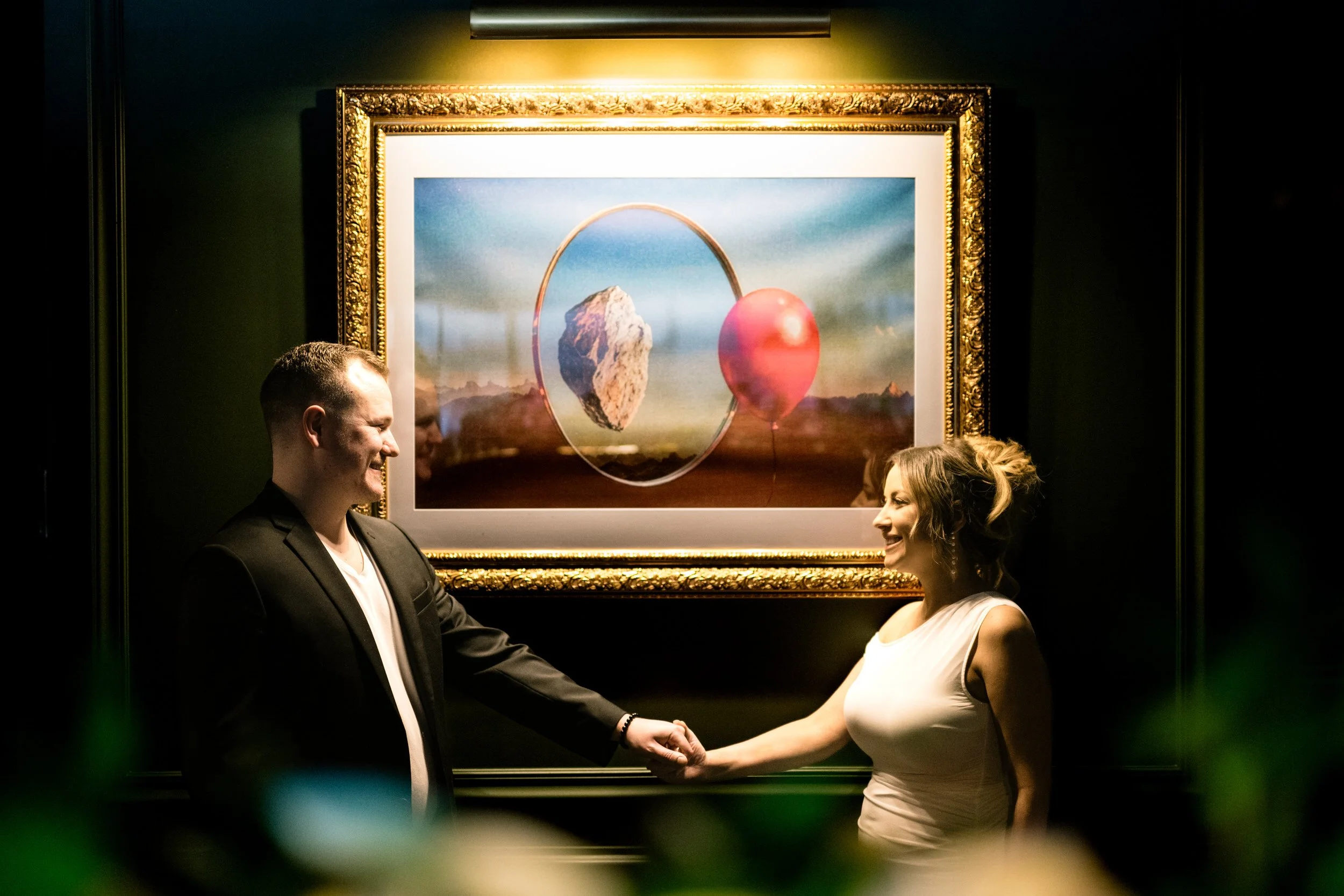 A couple holds hands with glowing light around them  and an artistic painting behind them.