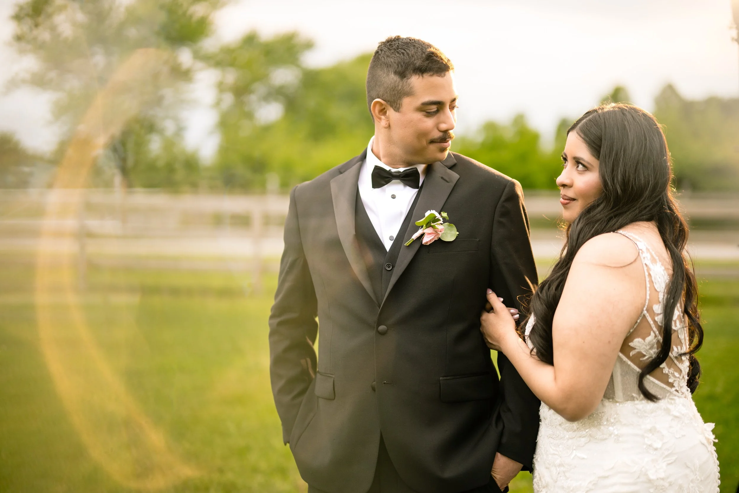 A man in a black tuxedo and a woman in a white wedding dress stand together outdoors on grass, with greenery and a soft light flare in the background, captured by a talented Northwest Indiana and Chicago wedding photographer.