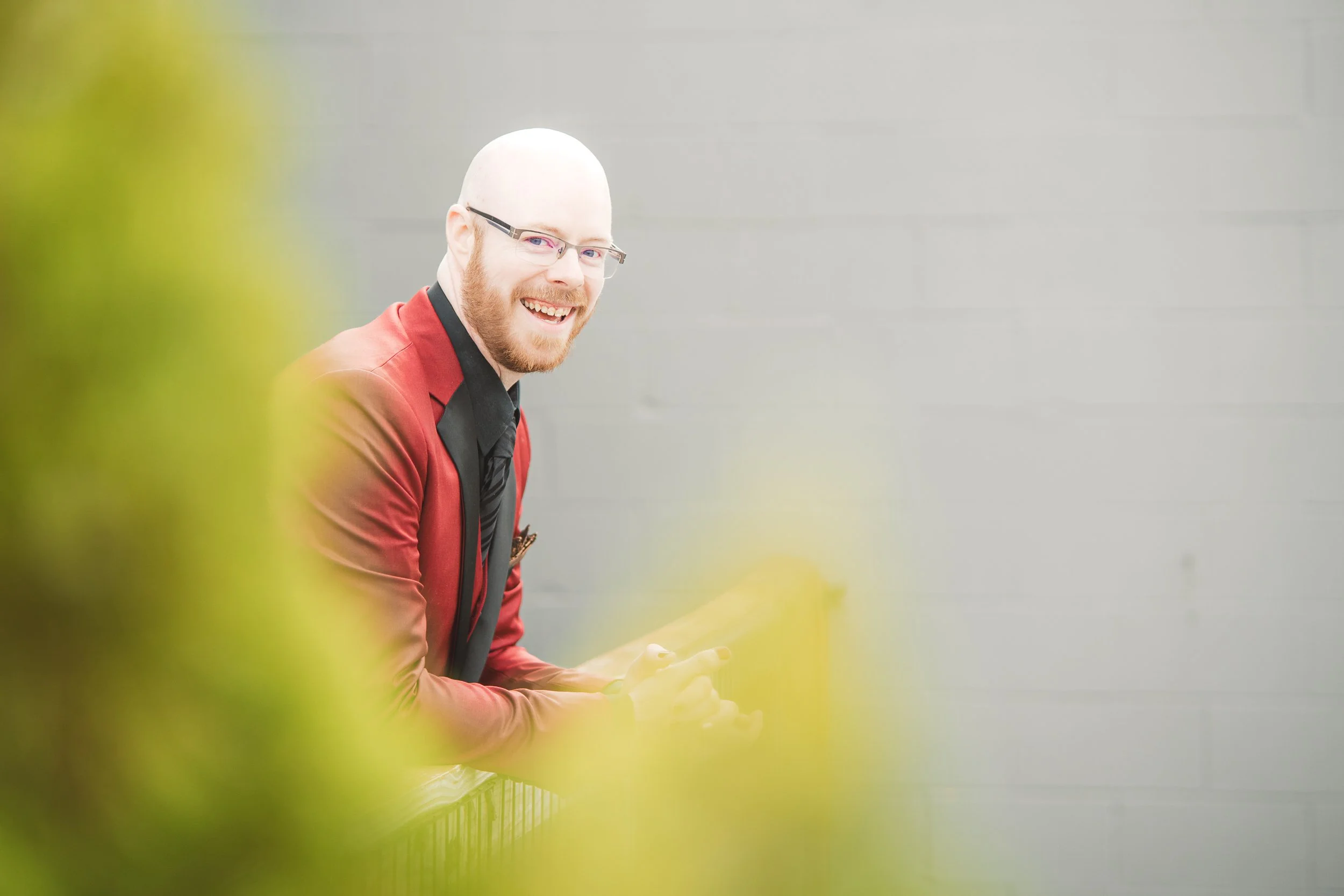 A groom laughs as he leans on railing and surrounded by greenery.
