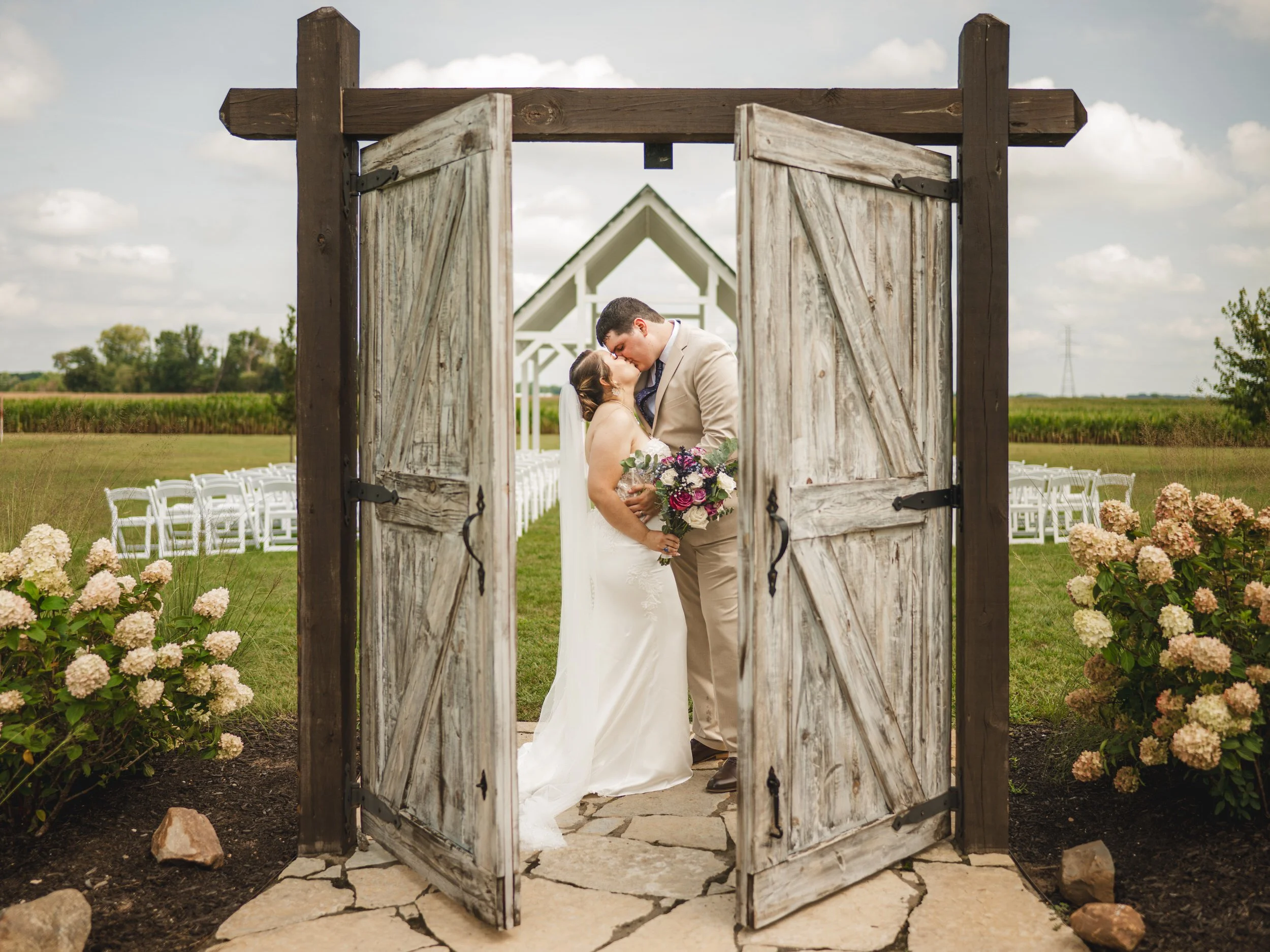 A bride and groom kiss under a rustic wooden archway at an outdoor wedding venue, beautifully captured by a northwest Indiana and Chicago wedding photographer, with white chairs and a small chapel in the background.