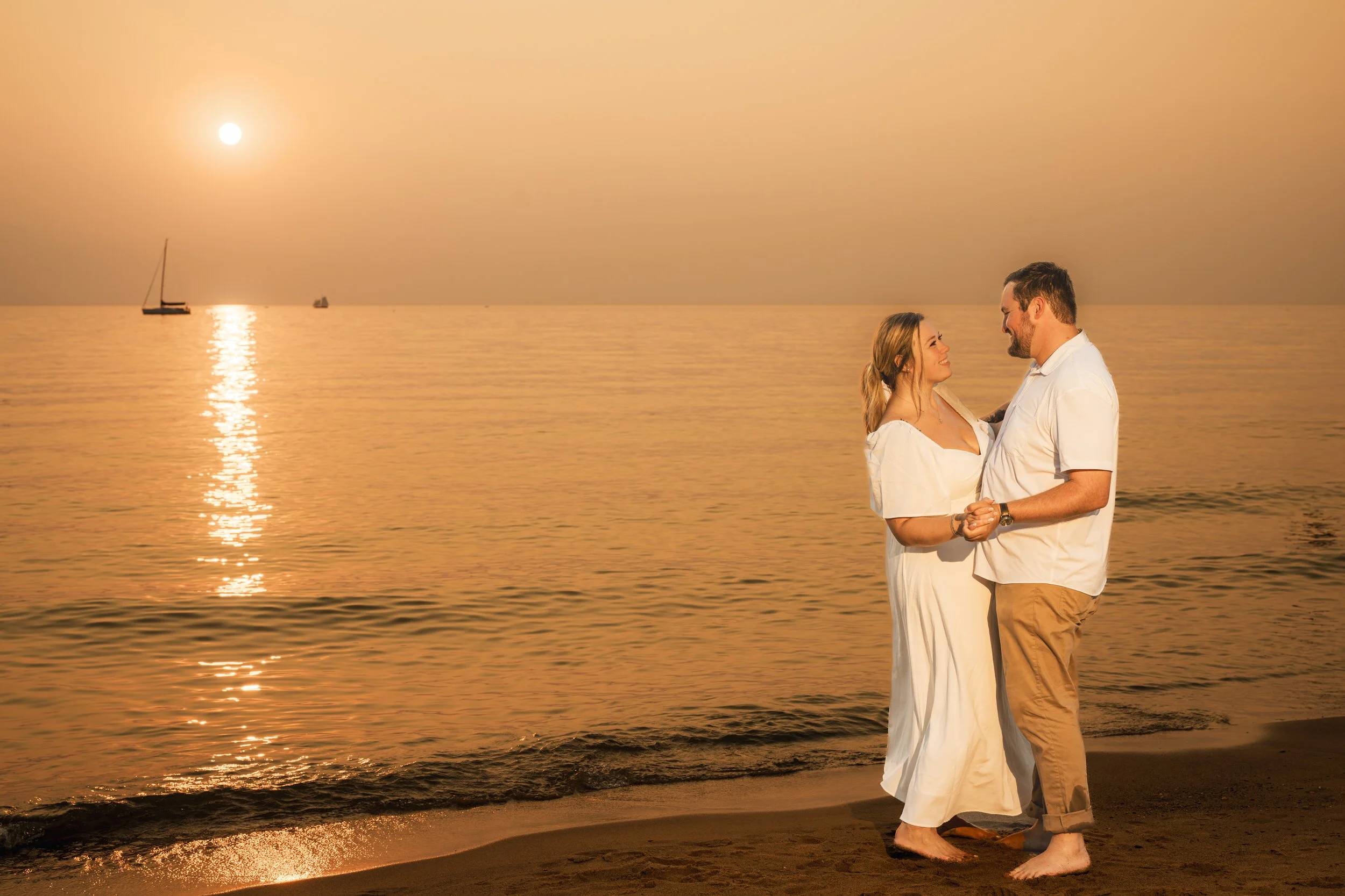 A couple stands barefoot on the sandy shore of Silver Lake Beach at golden hour, holding hands and smiling at each other as a sailboat drifts across the calm sea in the background, capturing the serene romance of southwest Michigan.