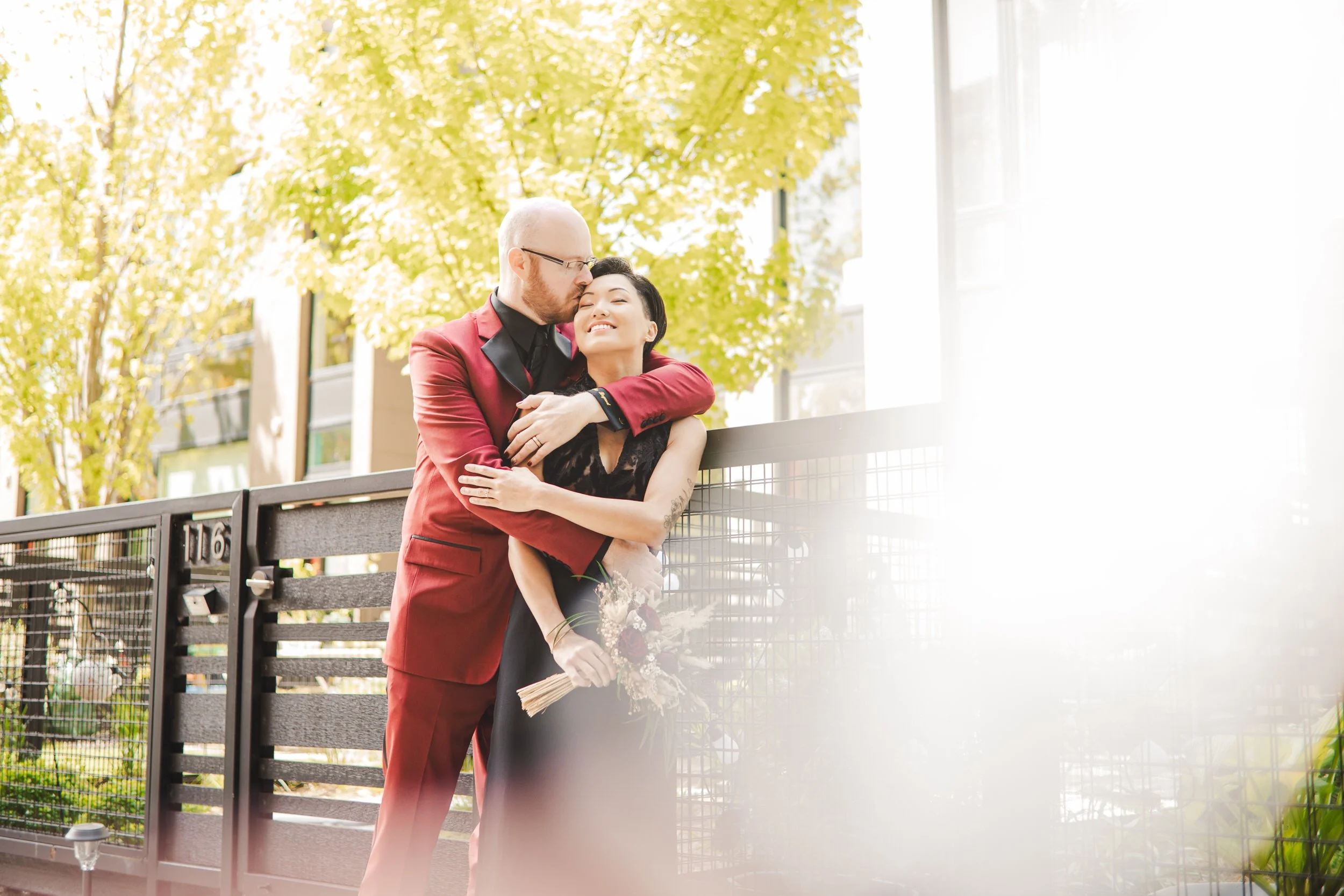 A groom in a maroon suit embraces his bride in a black lace weddingh dress among the greenery of a secret garden in Chicago.