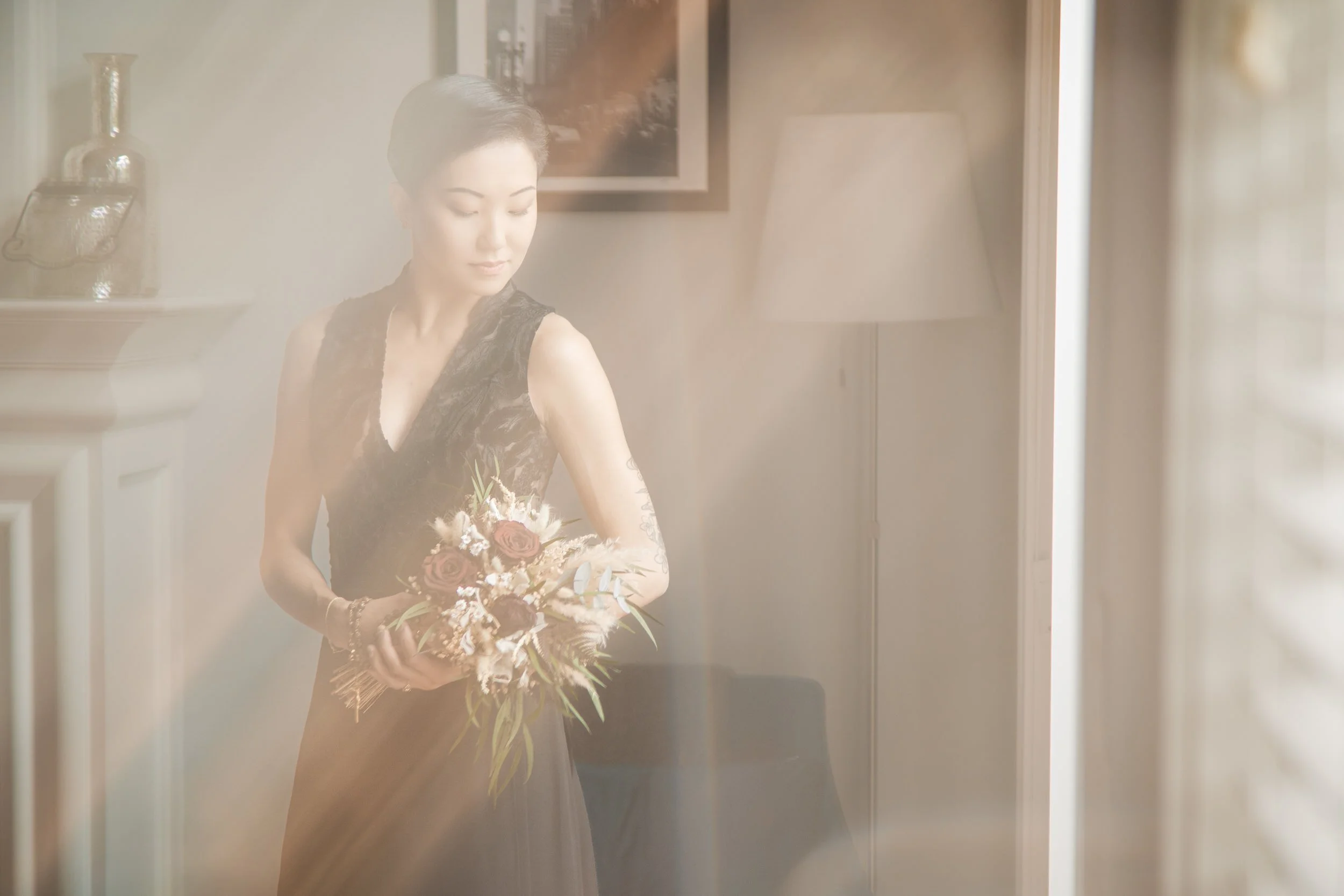 A beautiful Asian bride holding a bouquet of flowers as reflected light streams around her from an open window.