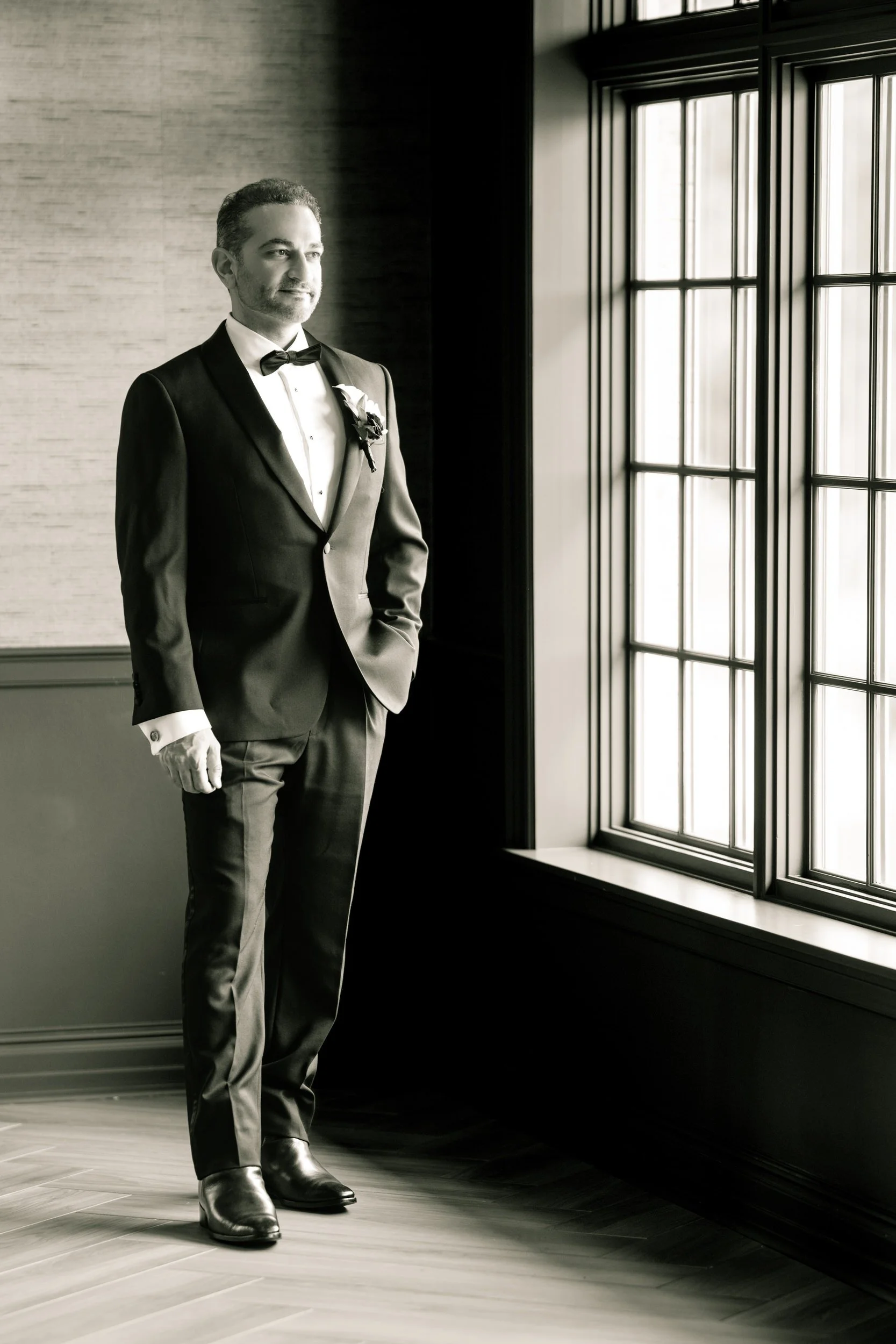 A black and white portrait of a groom looking outside the window at Midlothian Country Club before his outdoor wedding ceremony.