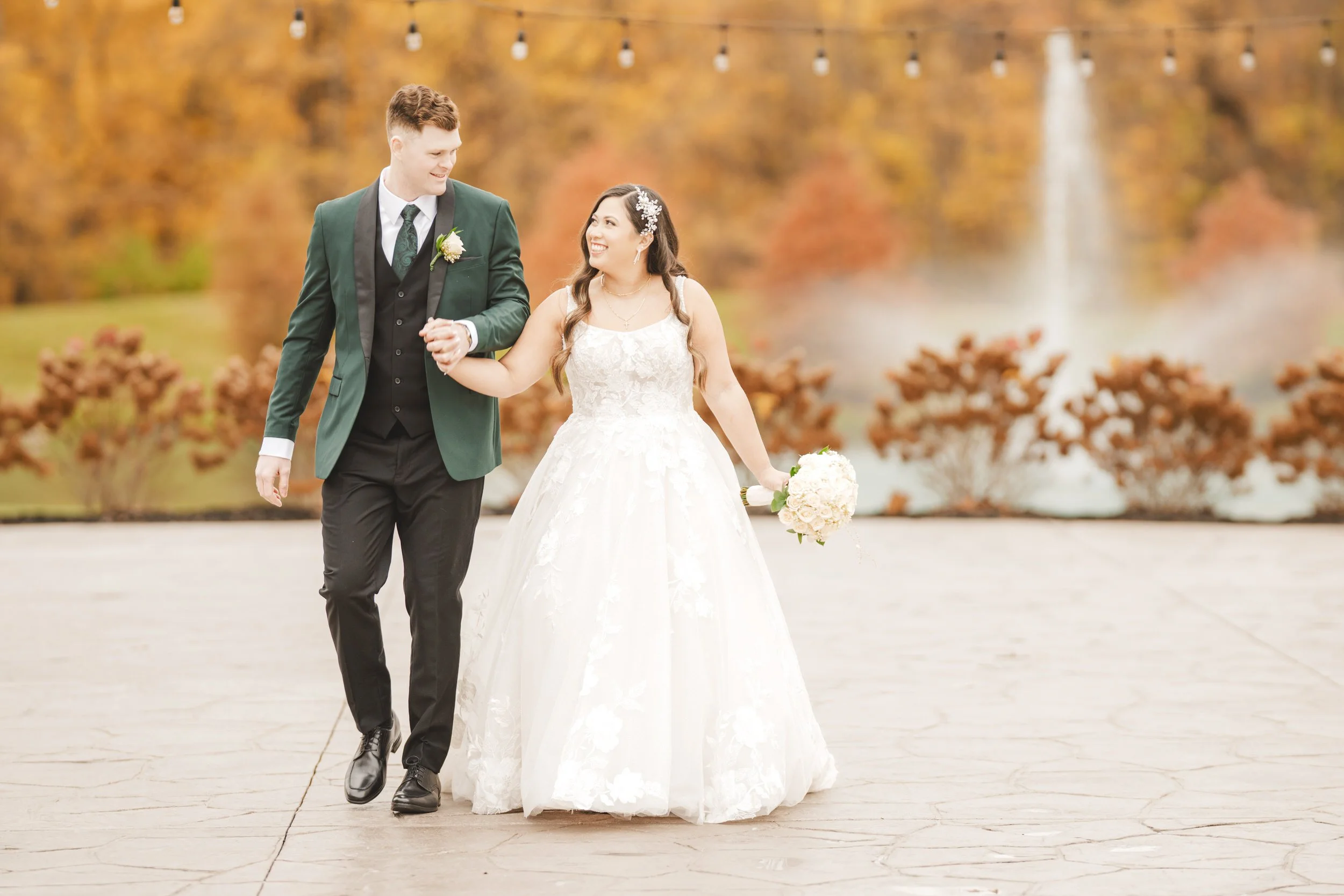 A bride and groom walk hand-in-hand outdoors, dressed in formal wedding attire, with autumn trees, string lights, and a fountain in the background—captured beautifully by a Northwest Indiana and Chicago wedding photographer.