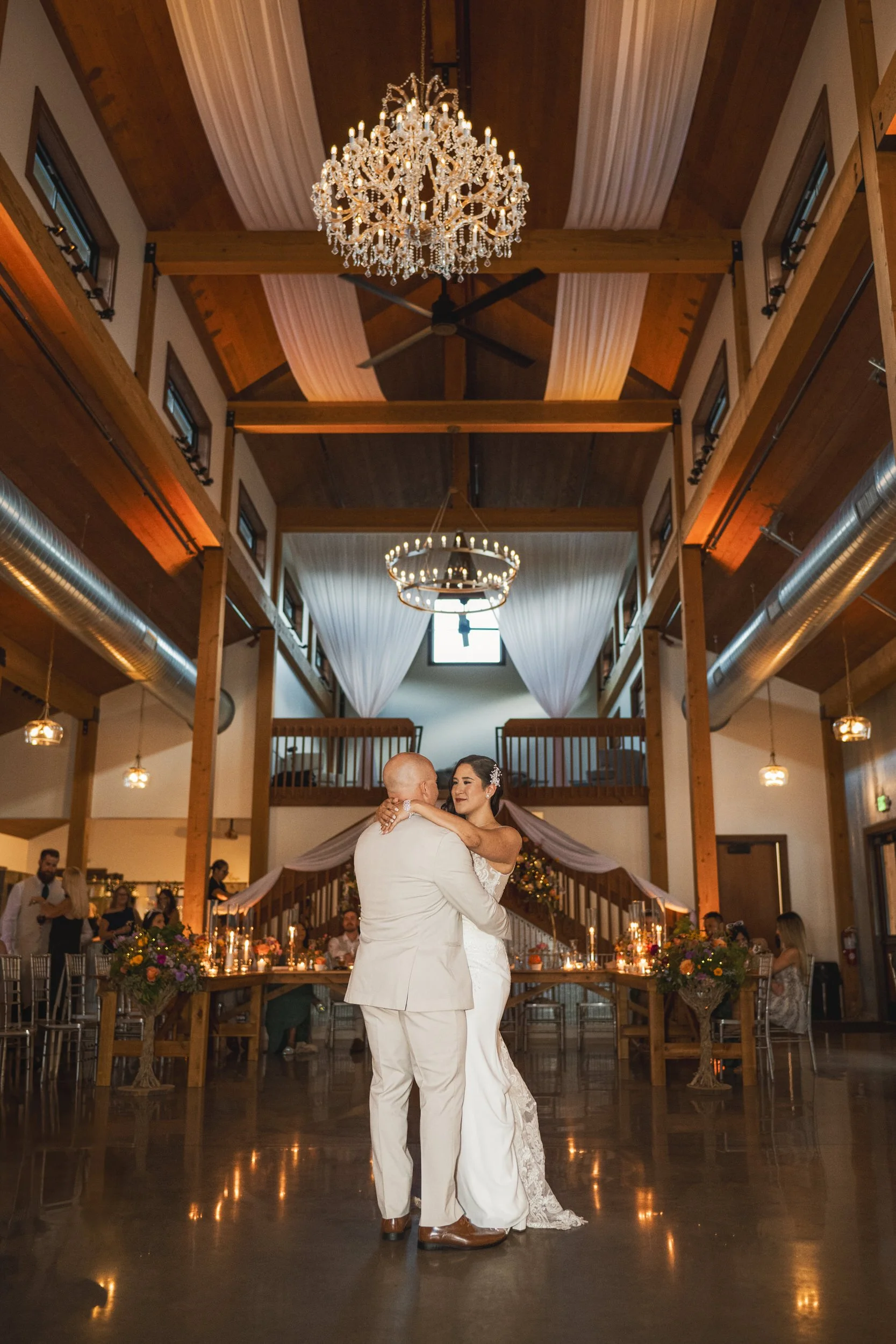 A bride and groom dance together in the center of a spacious, elegantly decorated hall with chandeliers and wooden beams overhead, captured beautifully by a northwest Indiana and Chicago wedding photographer.