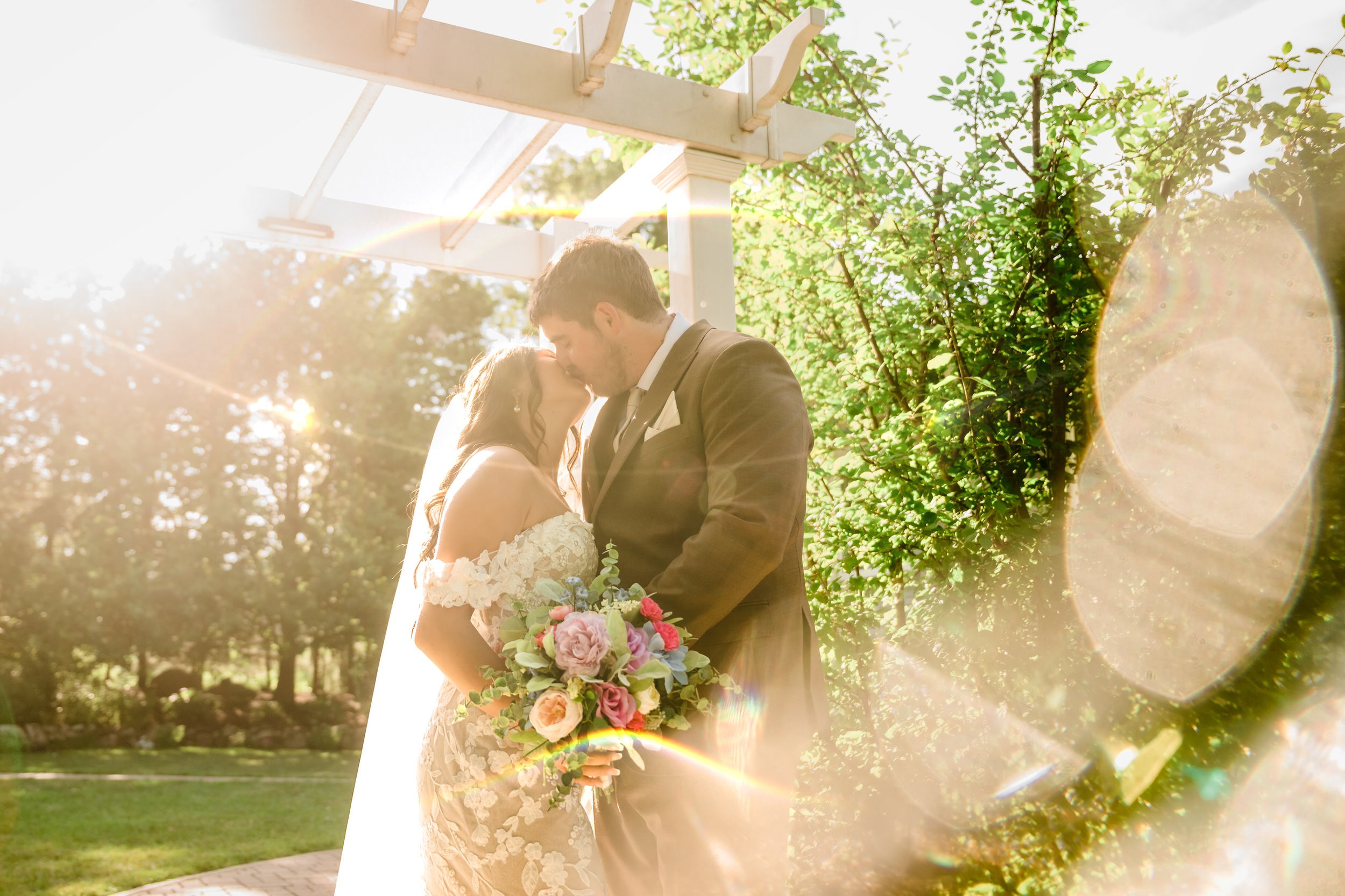 A bride and groom stand close together outdoors under a pergola, surrounded by greenery and sunlight, with the bride holding a colorful bouquet—captured by a Northwest Indiana and Chicago wedding photographer.