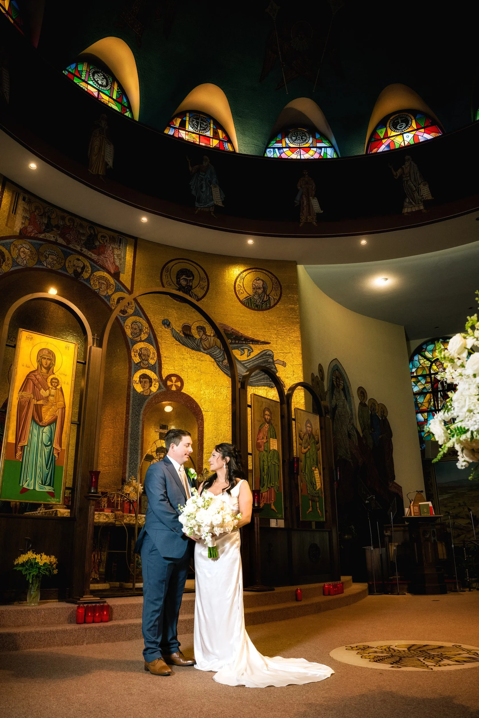 A bride and groom stand together holding flowers inside a church, surrounded by religious icons and stained glass windows, captured by a northwest Indiana and Chicago wedding photographer.